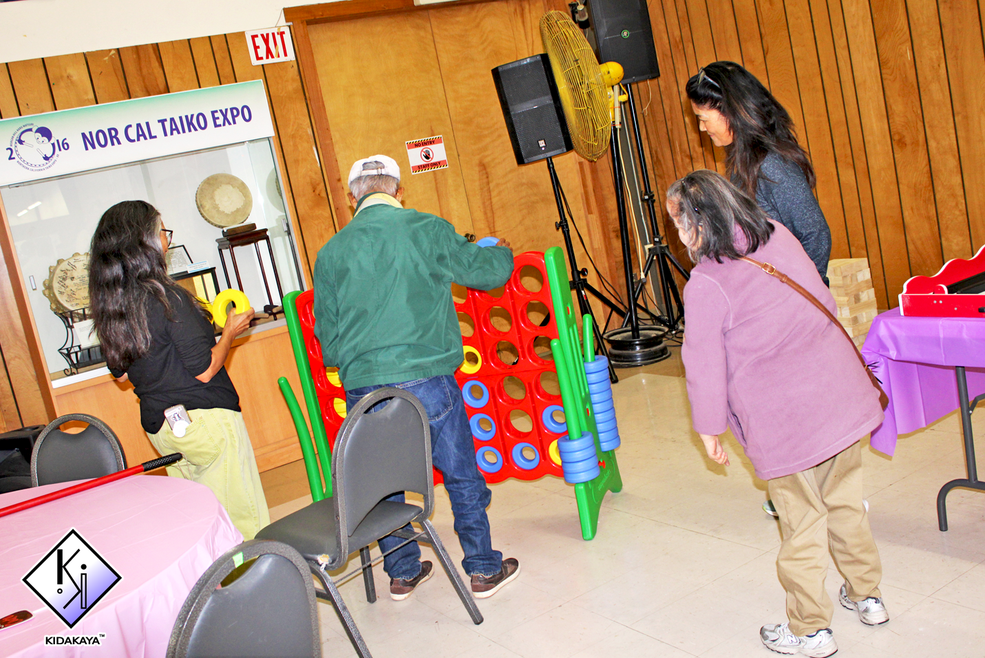 People playing a giant connect four game at the Nor Cal Taiko Expo, with display cases visible through an adjacent window and chairs surrounding pink and purple tables.