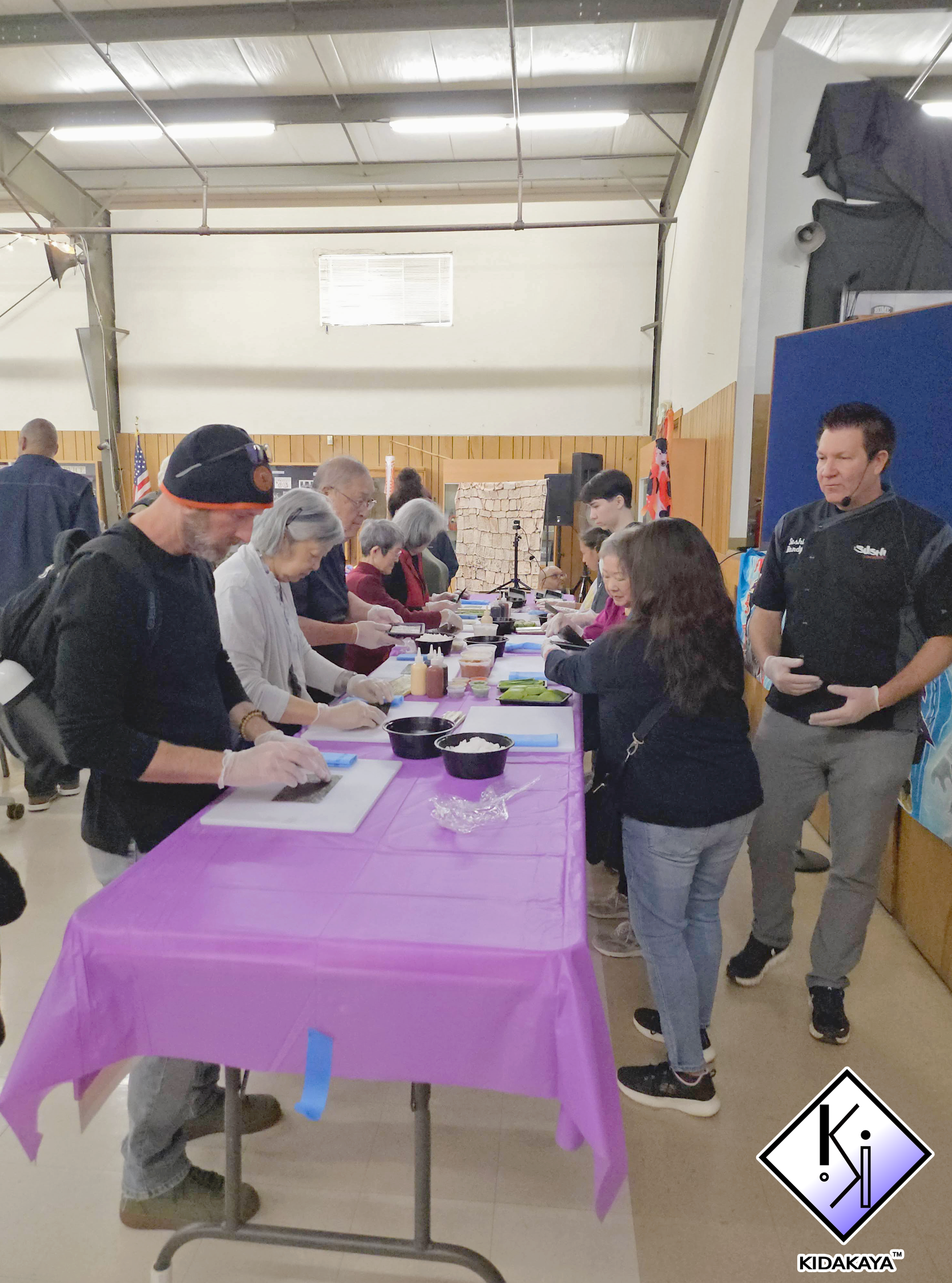 People participating in a community or craft event around a table covered with a purple tablecloth, with various supplies and ingredients, inside a community hall or church hall.