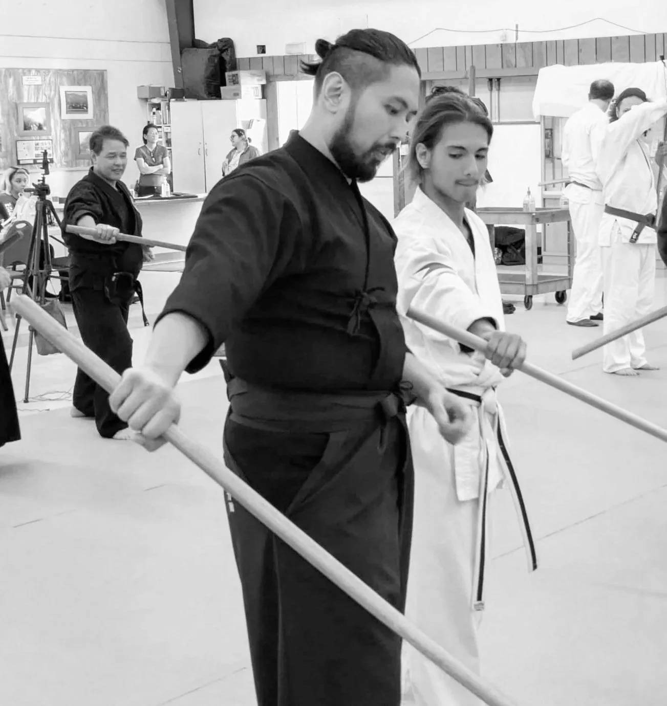 Two martial artists practicing in a dojo, one in a black gi and the other in a white gi, holding wooden swords.