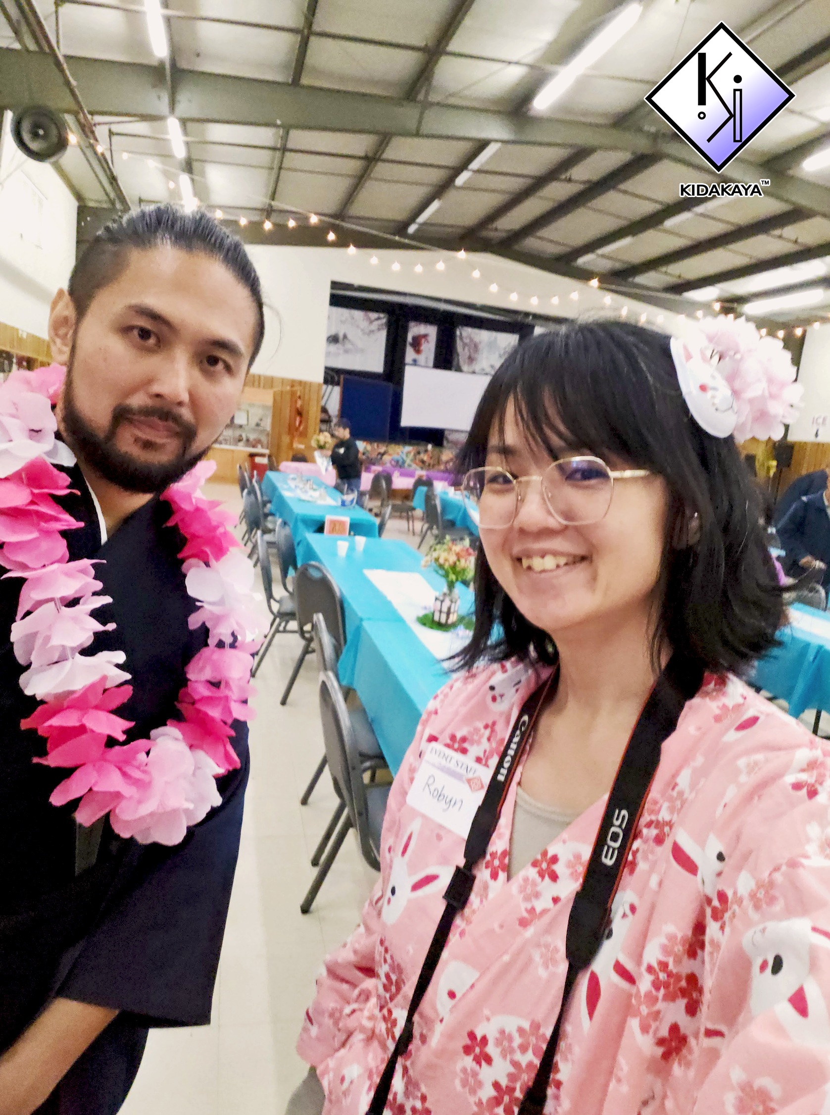 Two people taking a selfie at an indoor event with tables decorated with blue tablecloths and flower arrangements. The person on the left is wearing a black outfit and a pink flower lei, while the person on the right is wearing a pink floral kimono-s