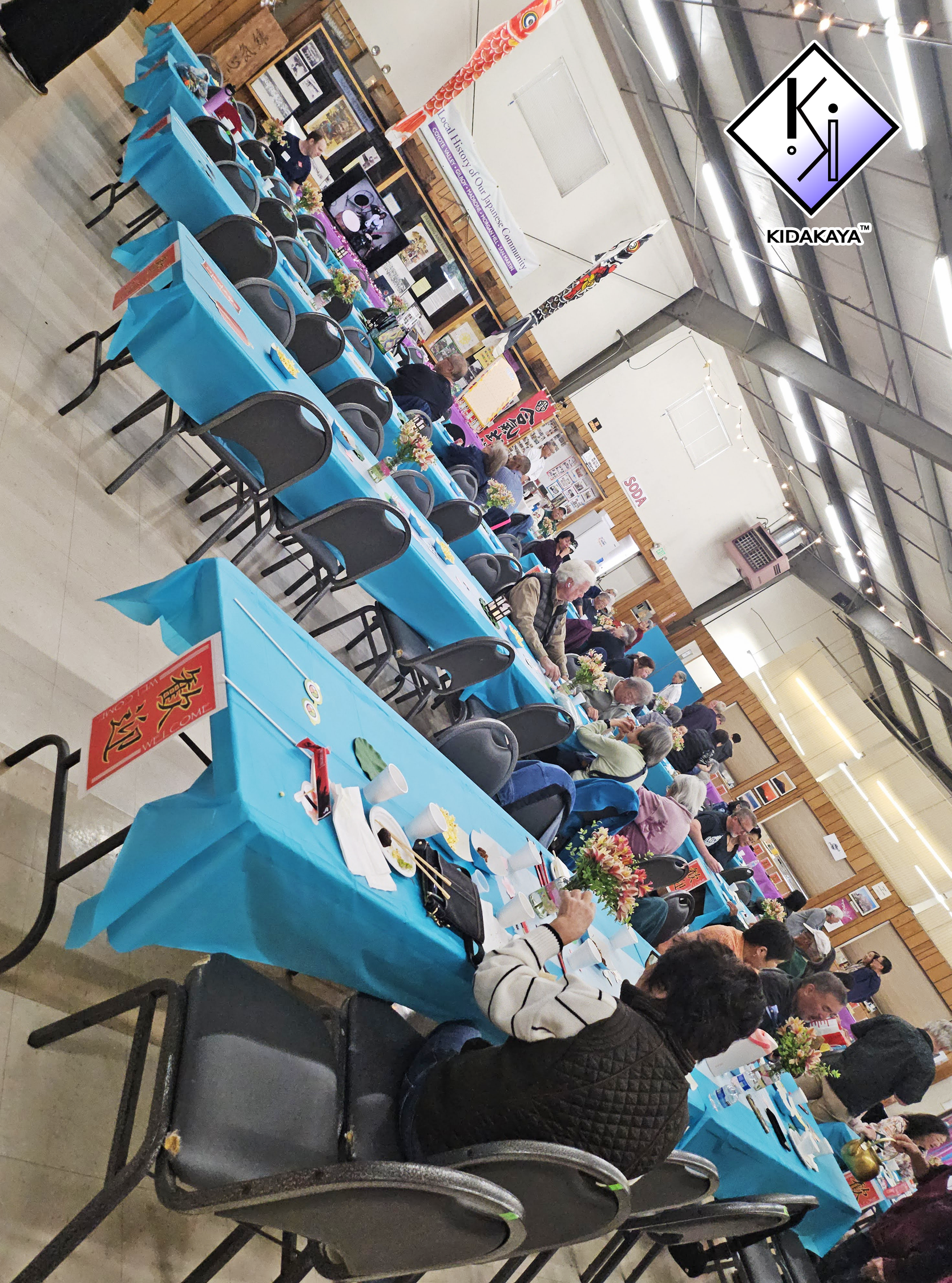 People sitting at chairs and blue tables decorated with flowers, dining and socializing in a large hall with display booths and banners, likely at a community event or cultural celebration.