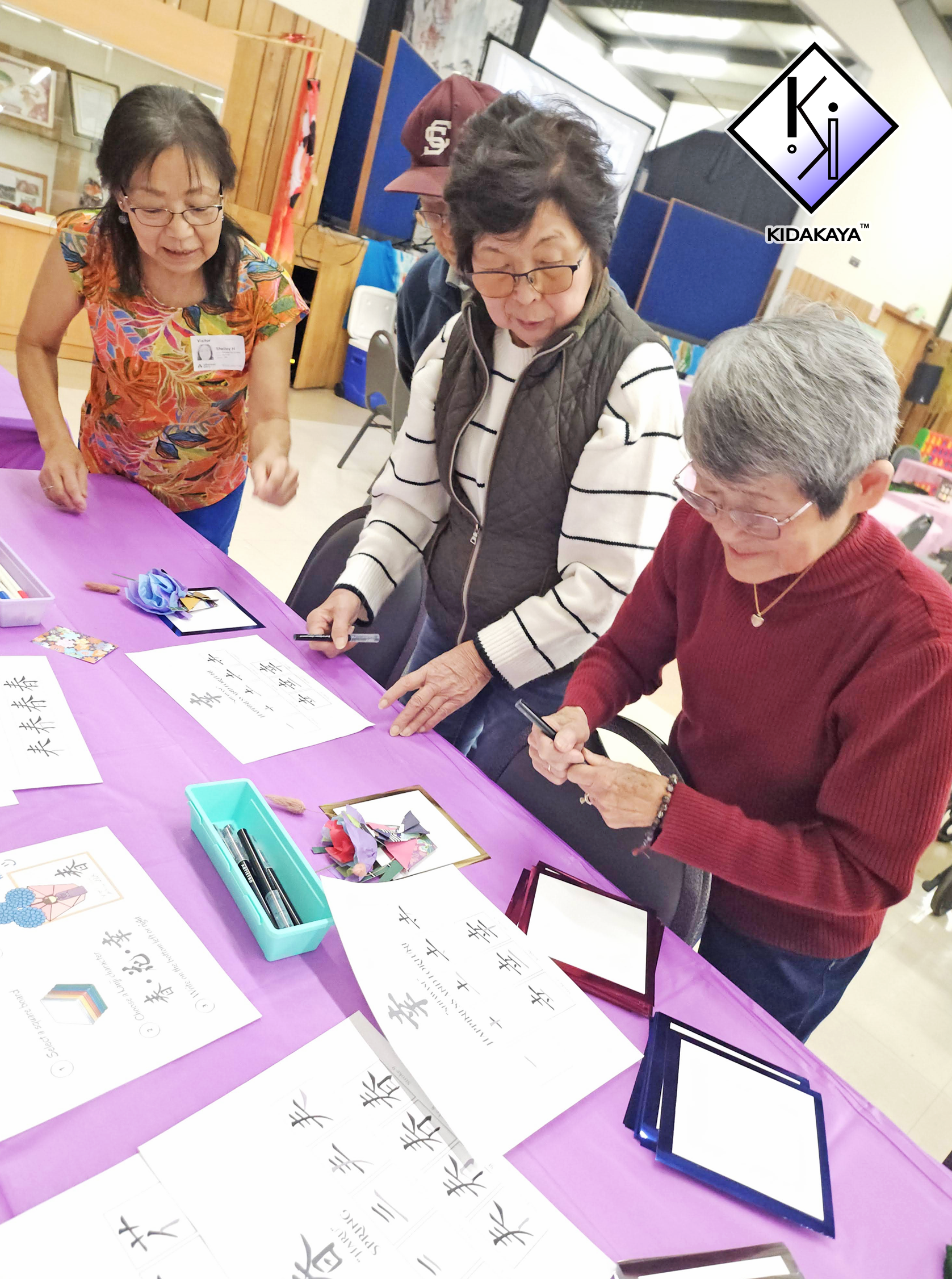 Four elderly women are gathered around a table with purple tablecloth, practicing Chinese calligraphy at a cultural event. The table has papers with Chinese characters, brushes, and decorative items. The background shows an indoor setting with wood p