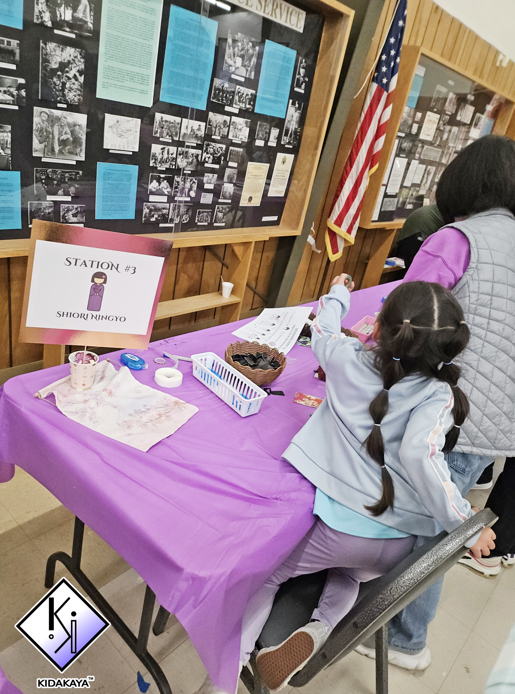 A table covered with a purple tablecloth with two children sitting and working on a craft project. Behind them, a display case with history photographs and blue informational sheets is visible, along with an American flag.
