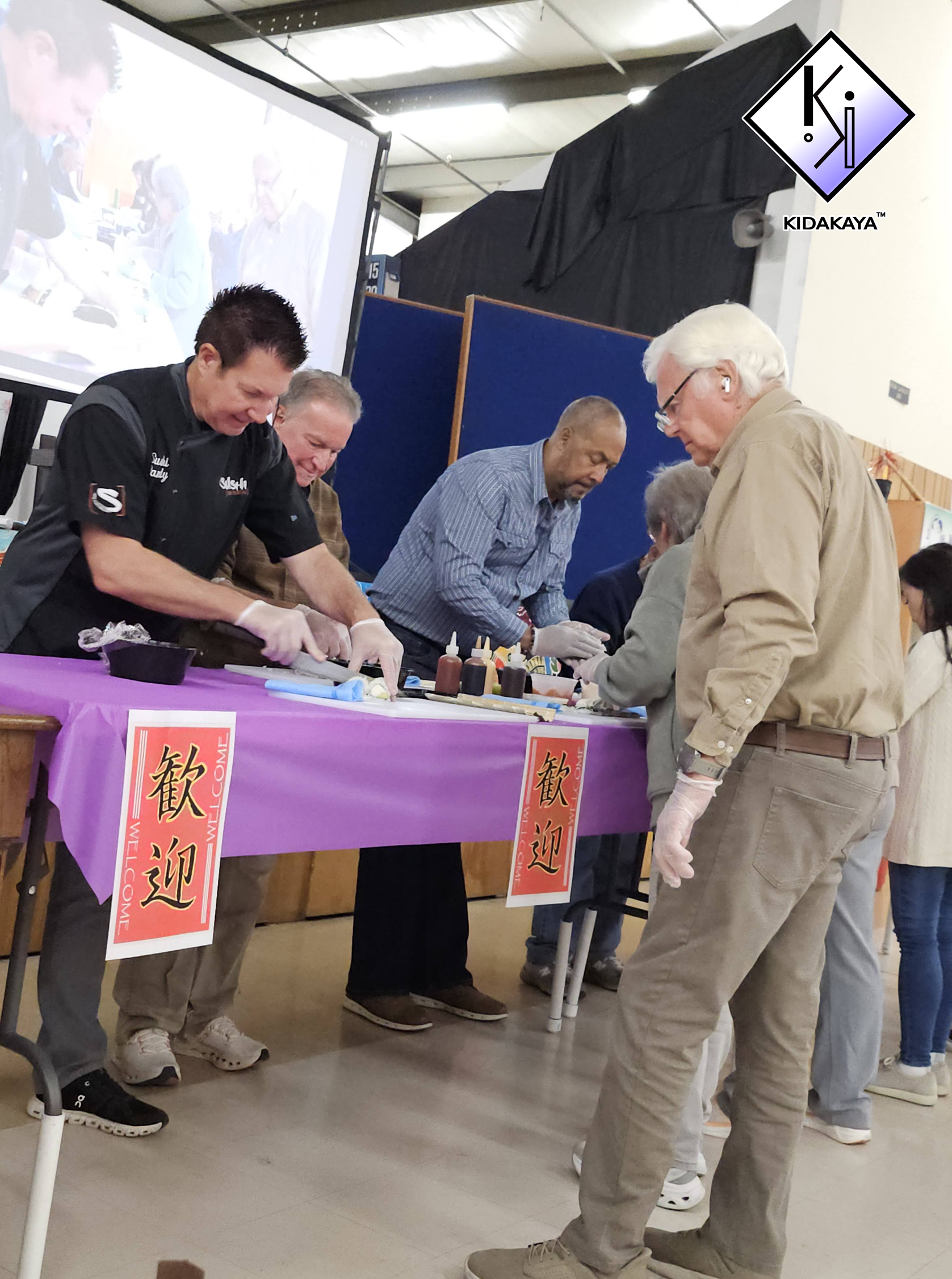 Several people at a table with a purple tablecloth, some wearing gloves, engaging in a food preparation activity. There are Chinese welcome signs hanging from the table. A backdrop and some display screens are visible in the background.
