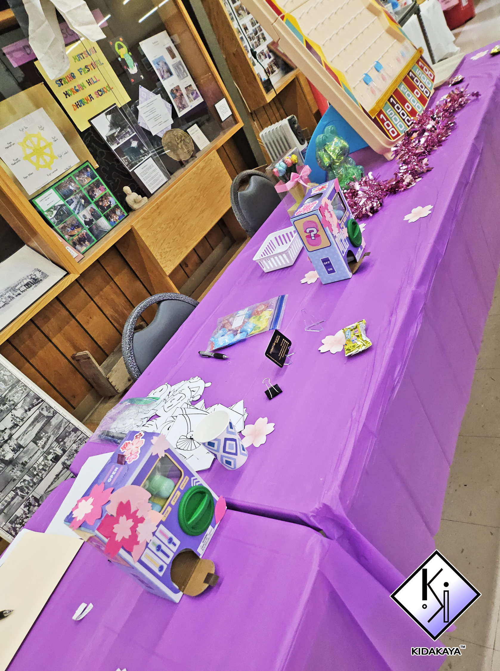 Table covered with purple tablecloth, decorated with pink tinsel, pink paper flowers, and various craft supplies including paper cutouts, a marker, and a stamp pad. In the background, there is a carnival-style game with a pink box and plastic prizes,