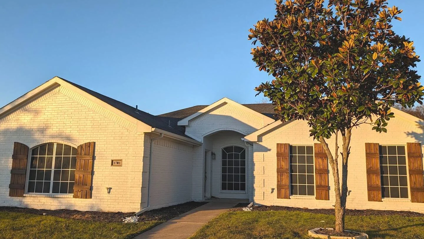 Front view of a white brick house with brown wooden shutters, a front door with an arch, and a small tree in the yard, during sunset.
