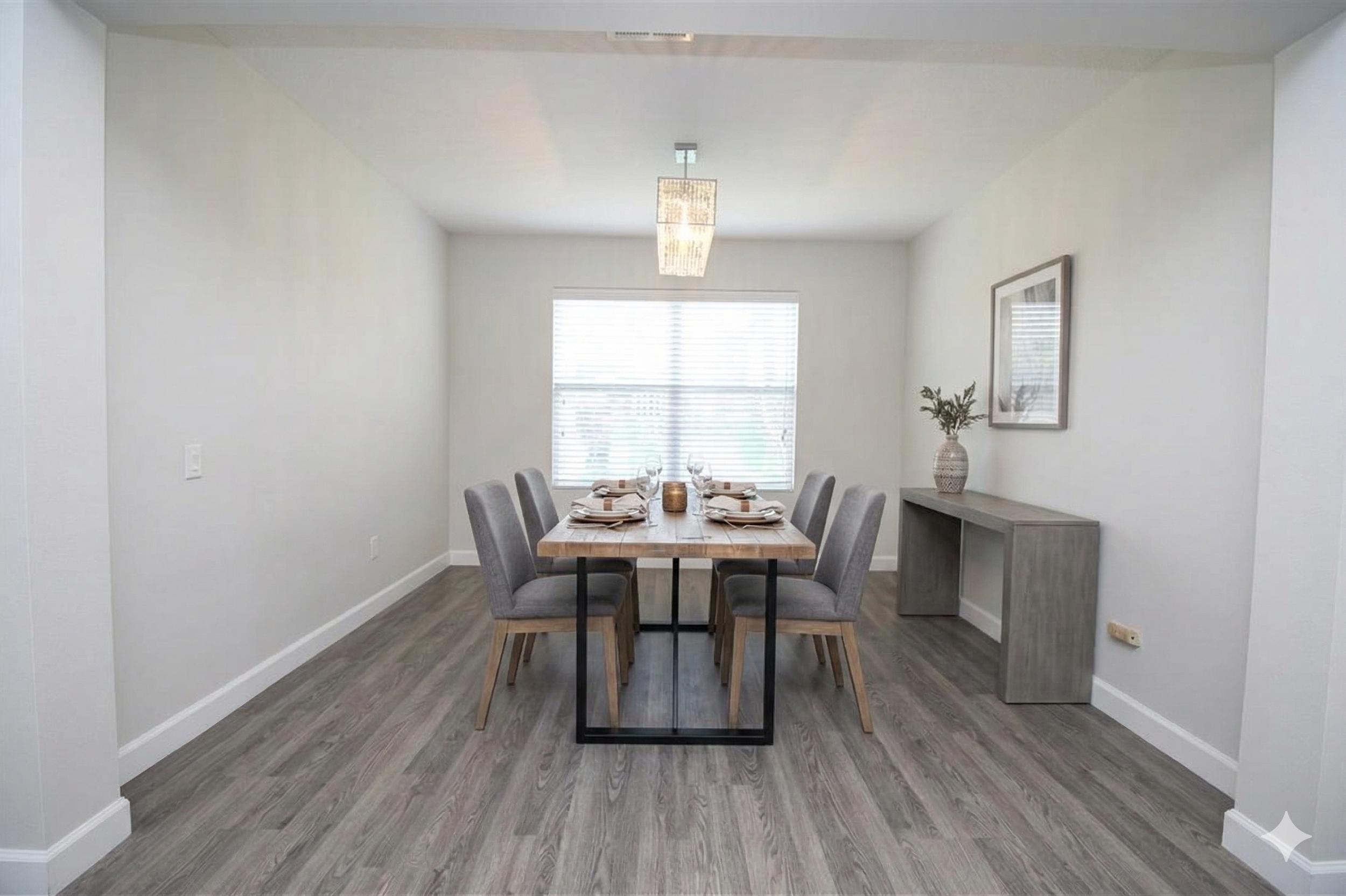 Modern dining room with a wooden table set for six, gray upholstered chairs, a large window with blinds, a chandelier, and a sideboard with a vase of greenery.