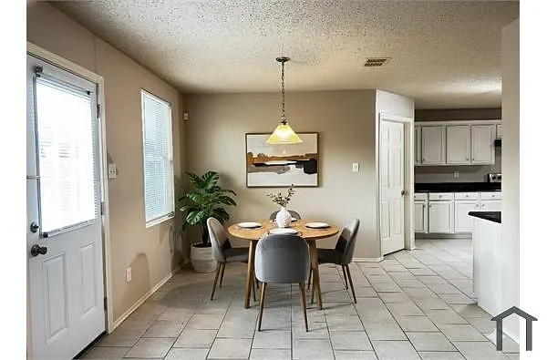 Dining area with a round table and four chairs, a green potted plant, a framed artwork, a hanging light fixture, and a view into the kitchen with white cabinets and tiled floor.