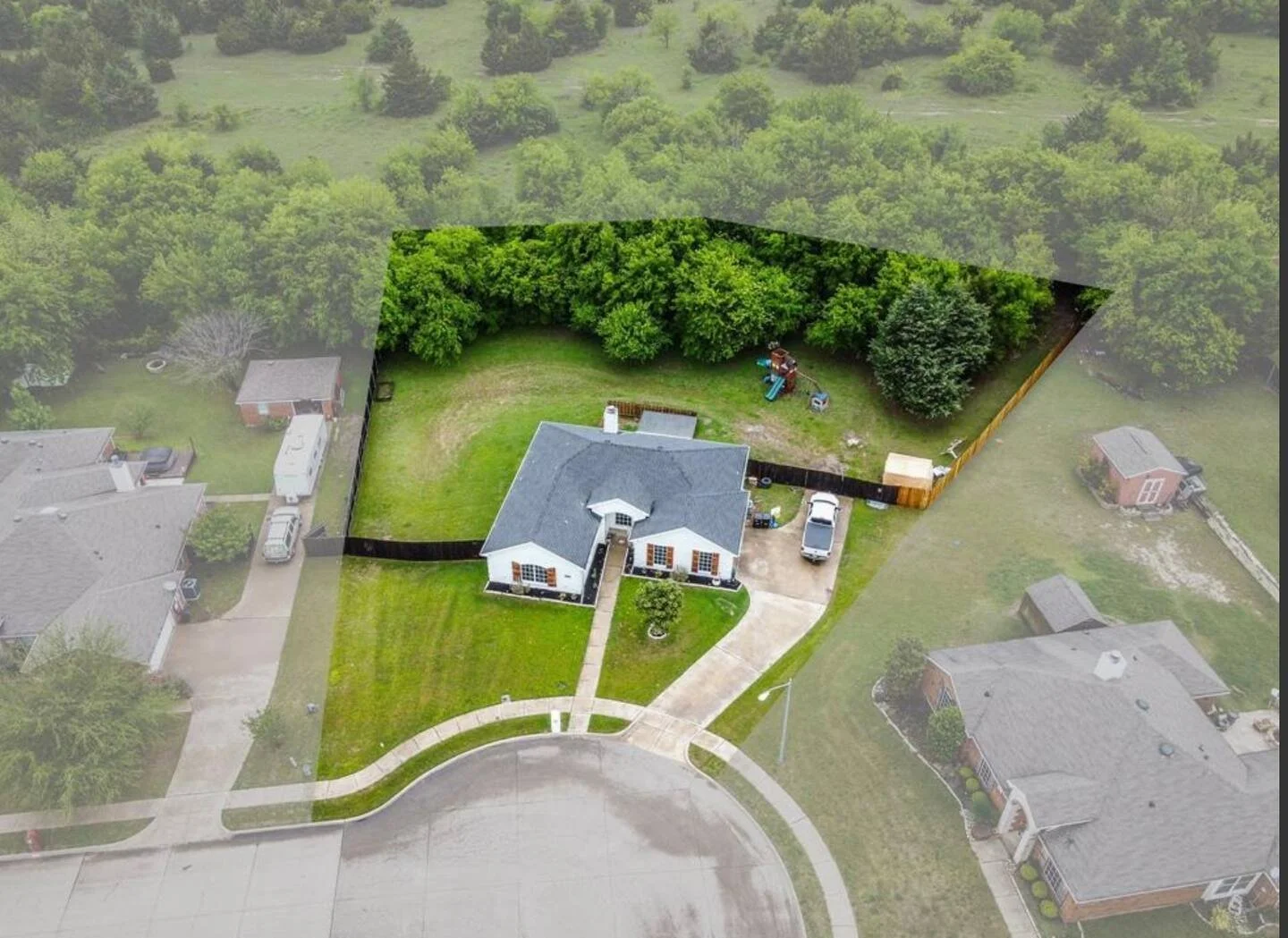 Aerial view of a house with a large backyard, partially fenced, with trees, a lawn, a driveway, a backyard playset, surrounded by neighboring houses and streets.