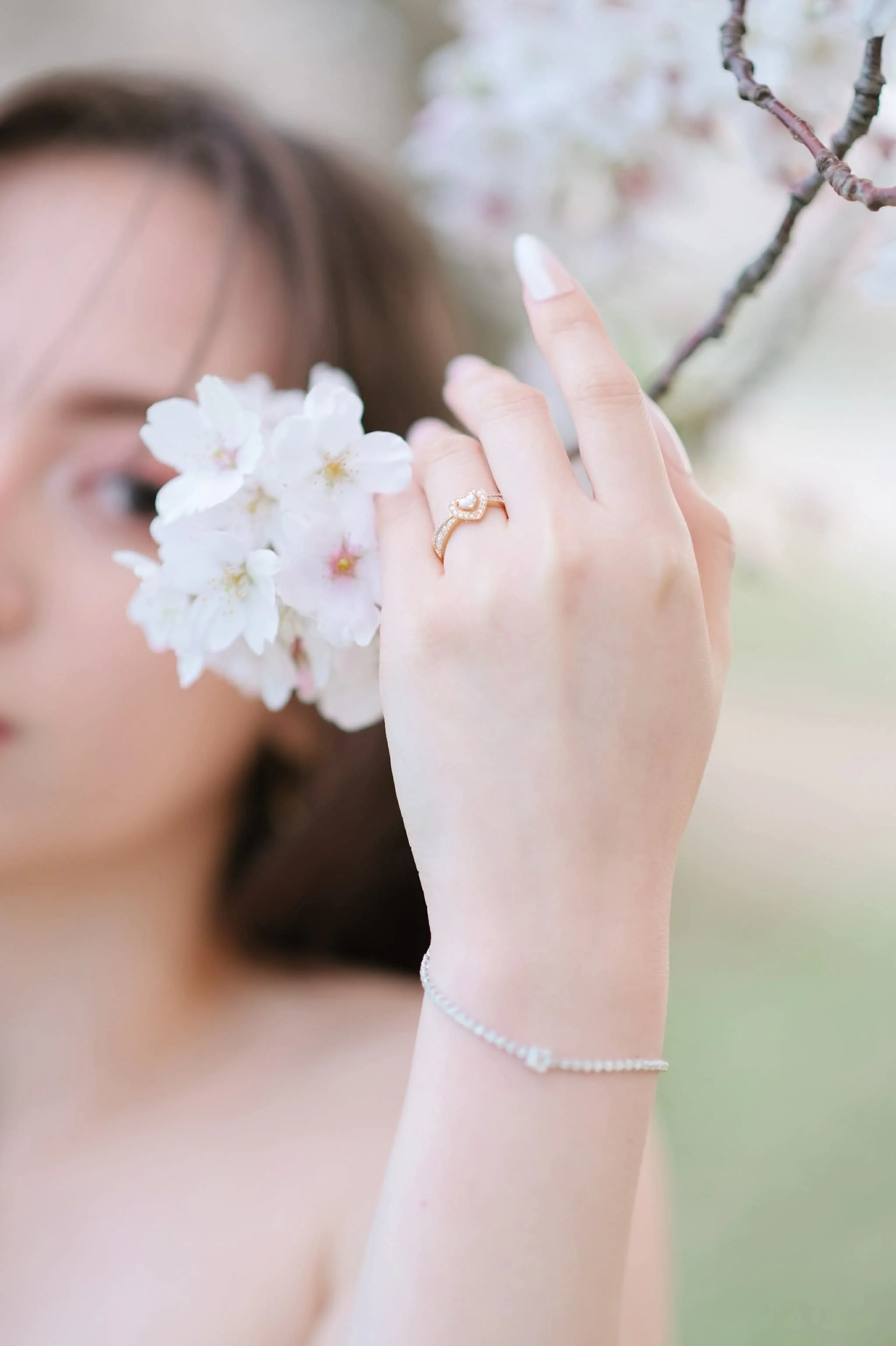 桜の花と女性の手のアップ。指にはハート型のダイヤモンドリング。背景はぼやけている。