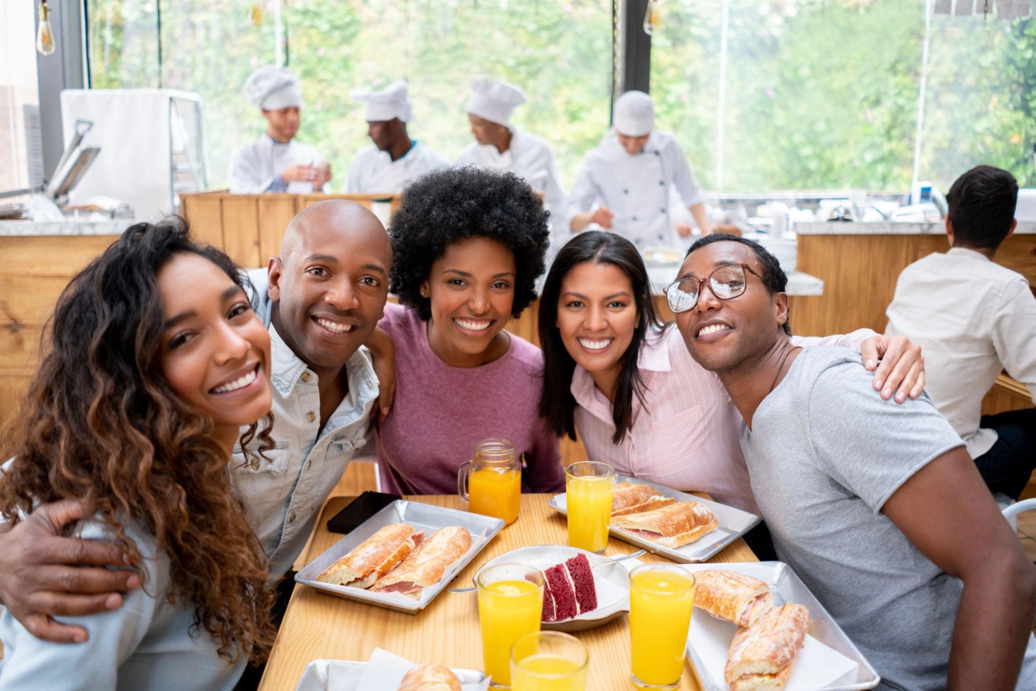 Group of five diverse people smiling, sitting at a breakfast table with sandwiches and glasses of orange juice, in a bright room with large windows in the background. Chefs are working in the kitchen area behind them.