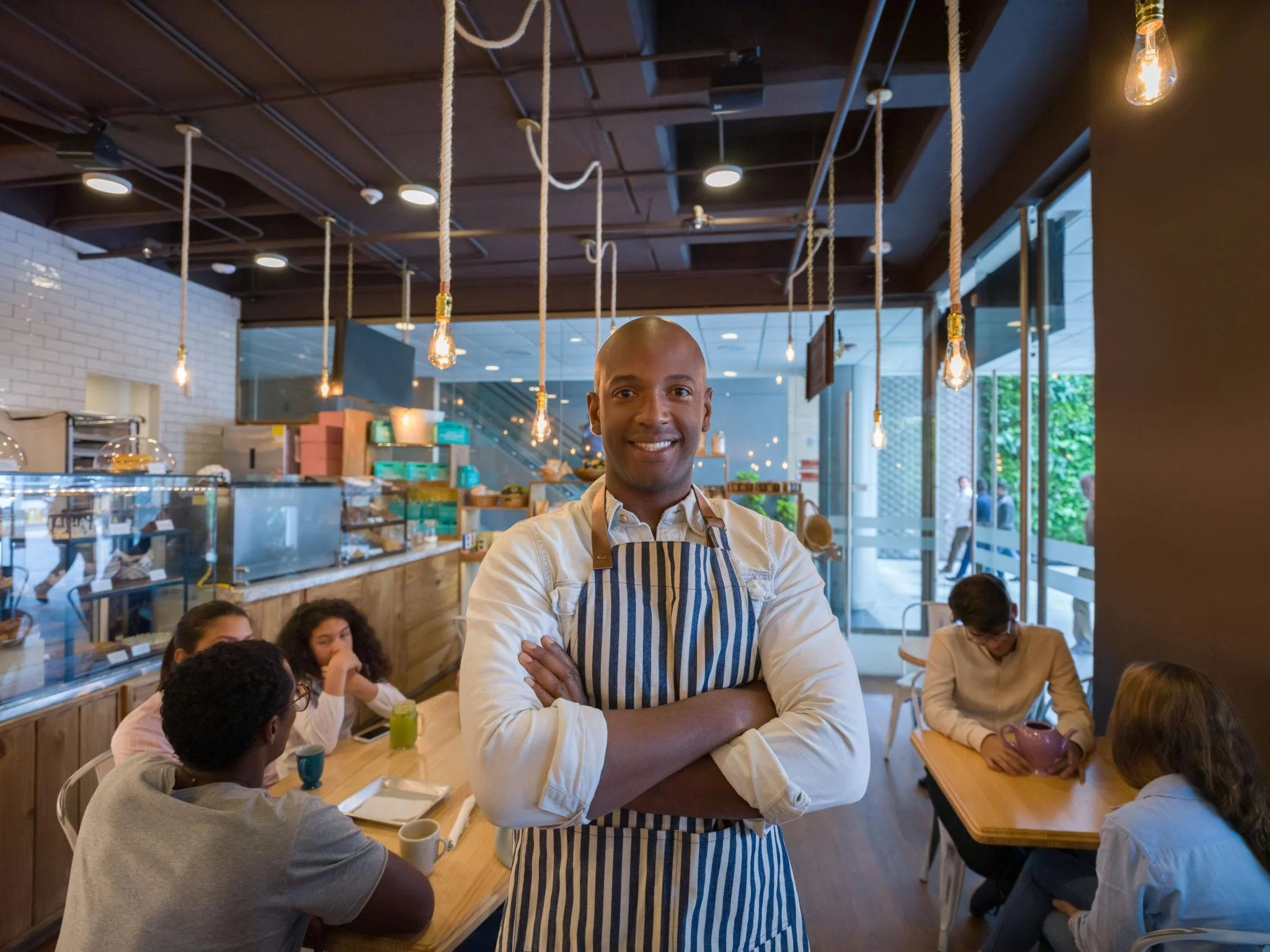 A smiling man with a shaved head wearing a striped apron stands with arms crossed inside a busy café or restaurant. Behind him, people are sitting at a long wooden table, some with coffee mugs and drinks. The café features modern lighting with hanging Edison bulbs, a white brick wall, and large windows showing outdoor greenery.