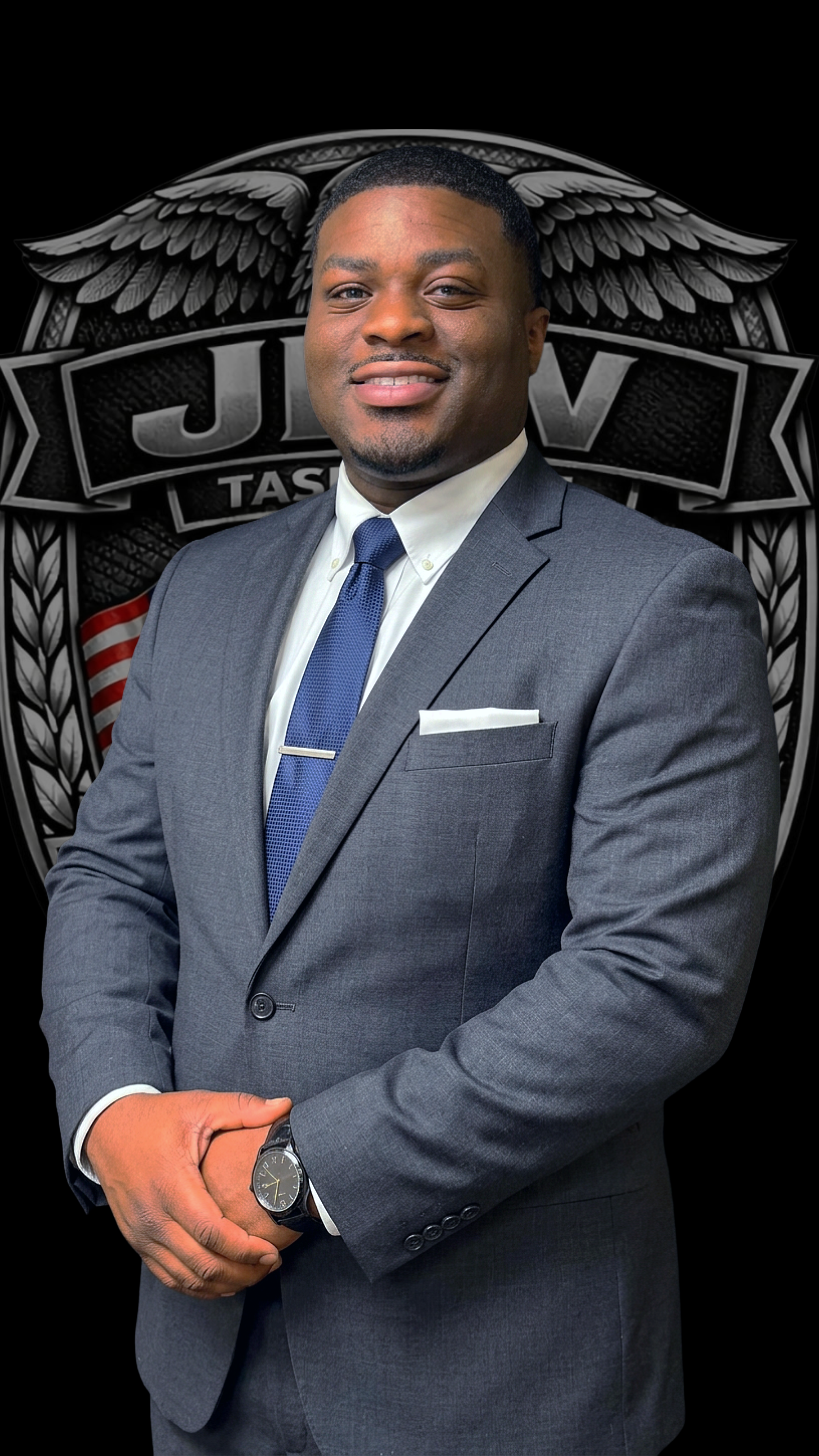 A man in a gray business suit with a white shirt and blue tie, wearing a watch, stands in front of a black background with a large logo that has wings and the words "J.U.V." and "Tactical".