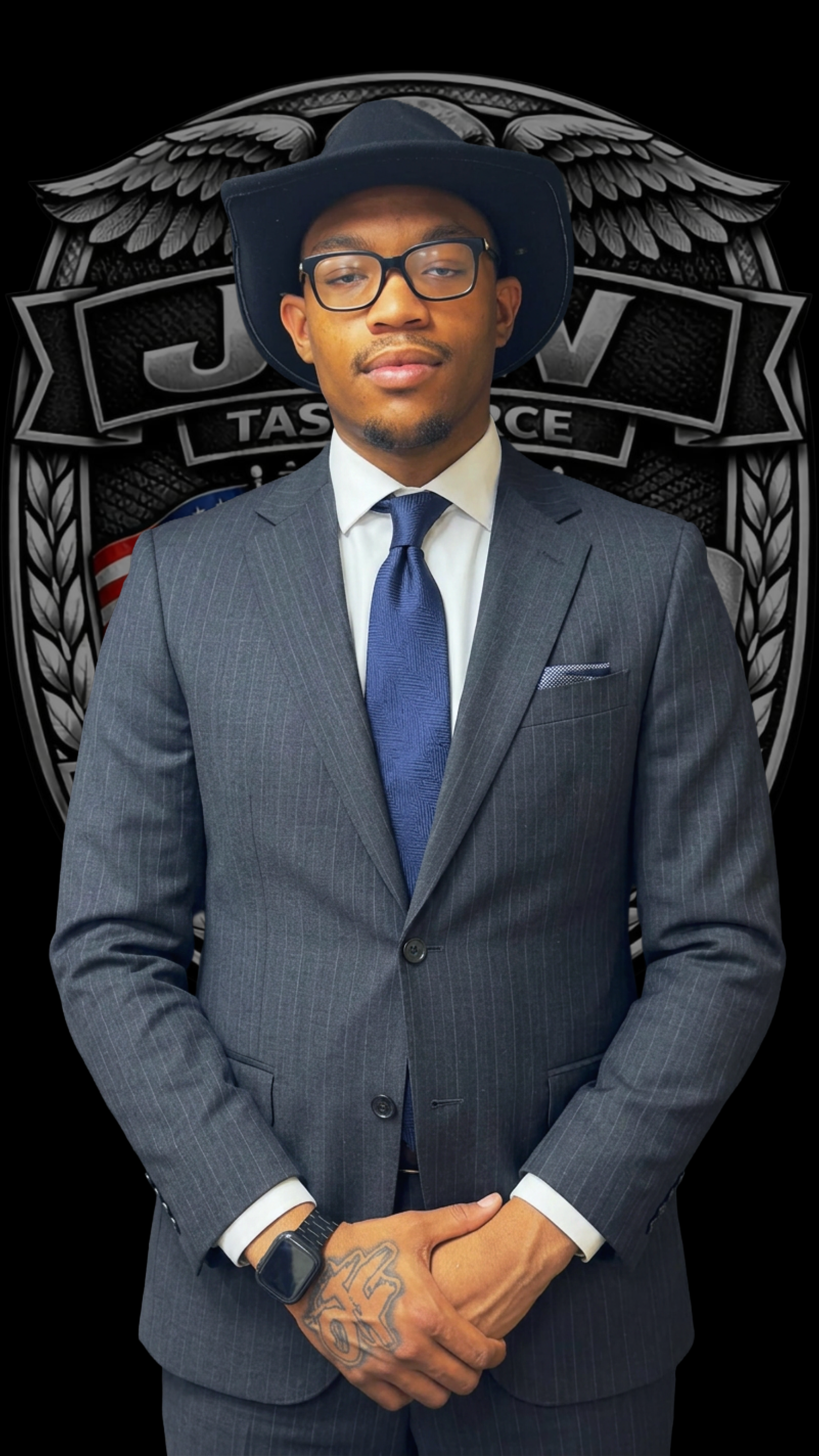 A man wearing a suit, blue tie, glasses, and a wide-brimmed black hat stands in front of a black badge with wings and the words "JDM Task Force" behind him.