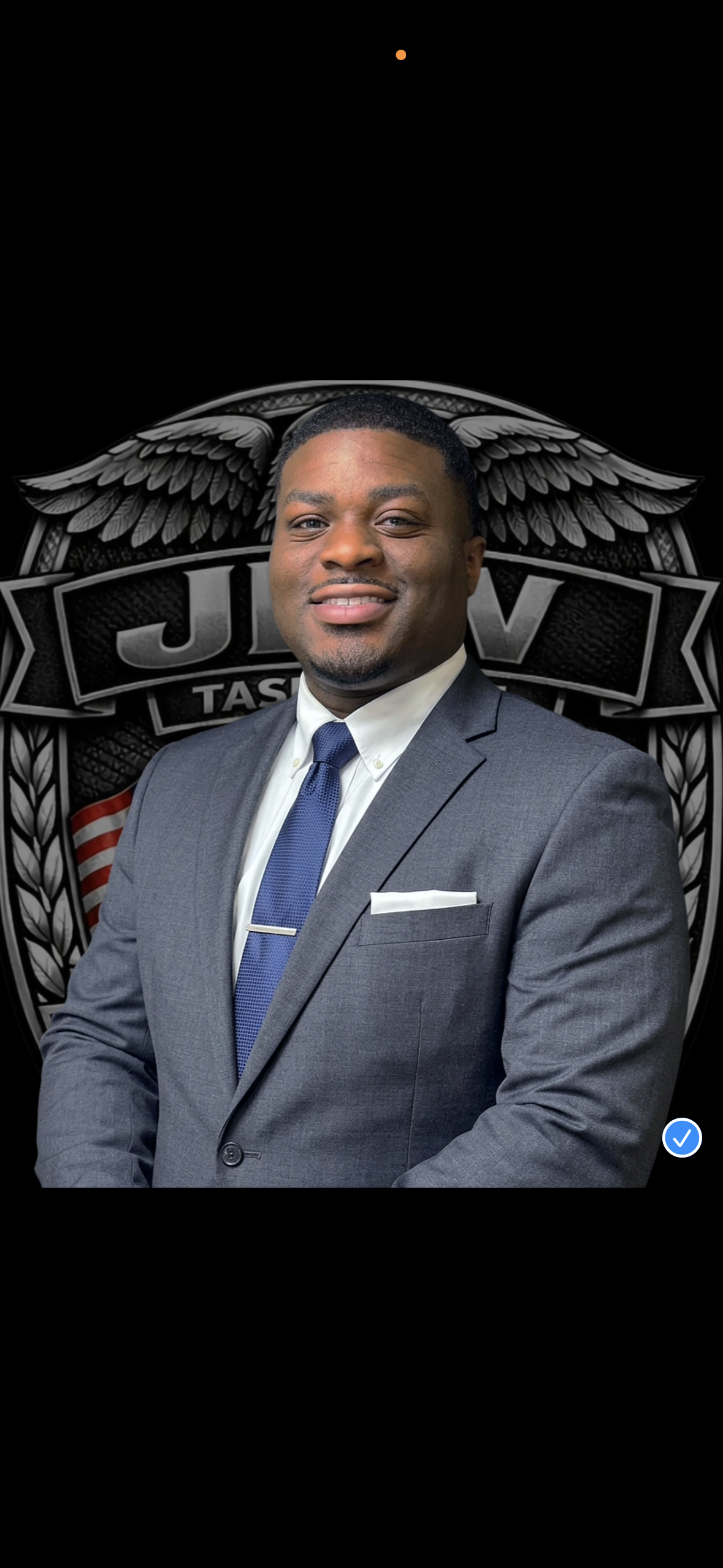 A smiling man in a gray suit and blue tie standing in front of a black logo with the word 'JAV' and eagle wings.