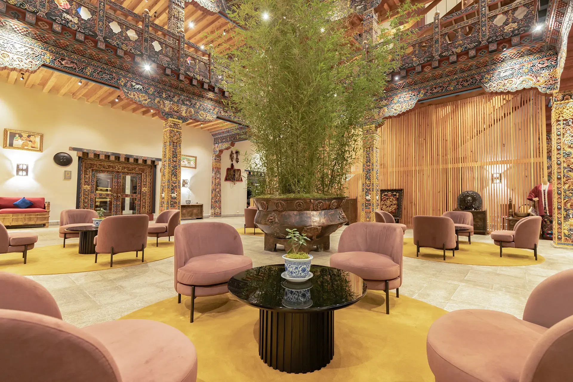 An elegant hotel lobby with traditional Asian interior design, featuring a large central potted bamboo plant, pink cushioned chairs arranged in a circle around black marble tables, intricate woodwork and decorative elements on the walls and ceiling, and various Asian-inspired sculptures and artwork.
