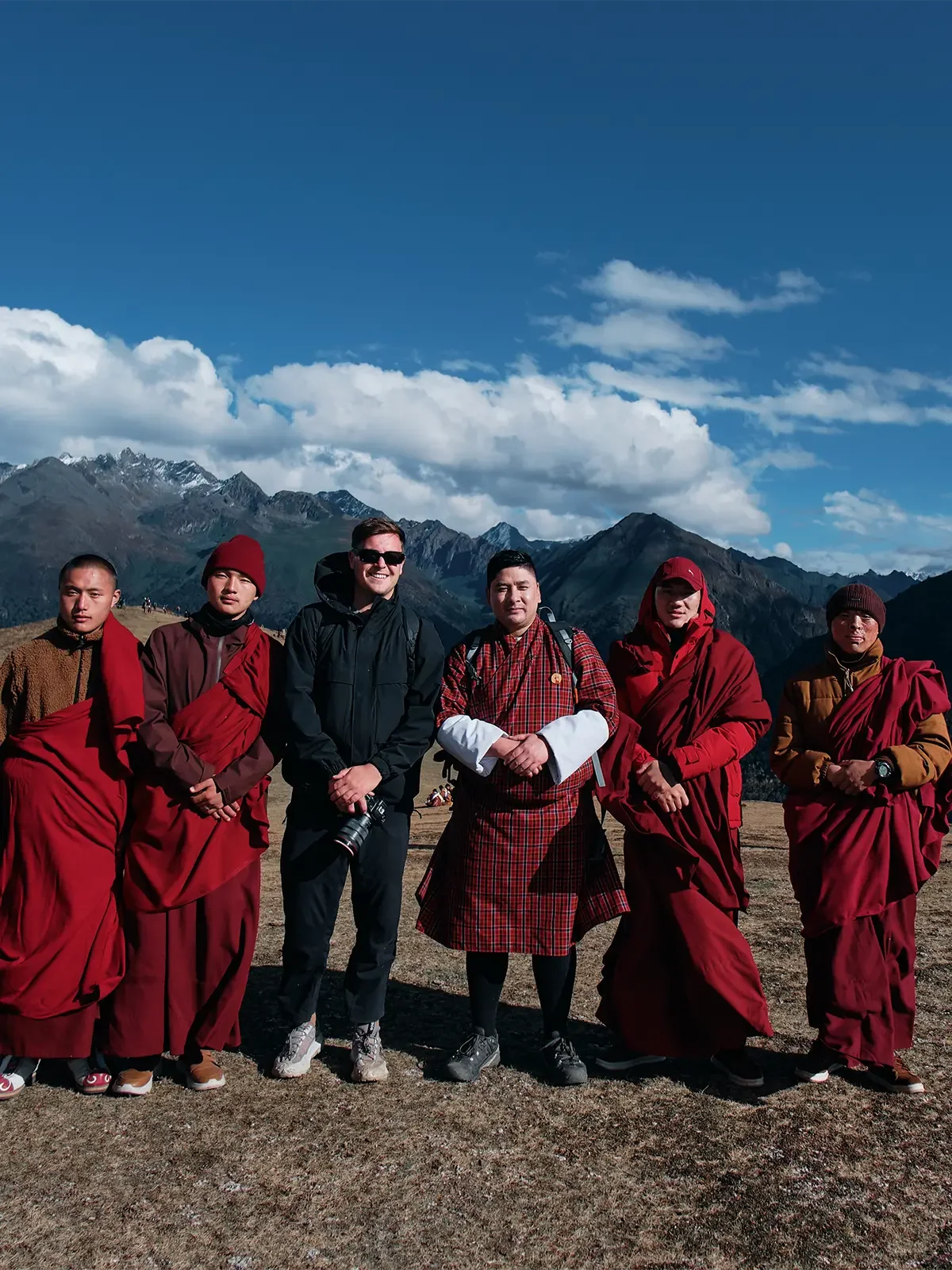 Group of tourists and monks standing together outdoors against a mountain landscape with a blue sky and clouds.