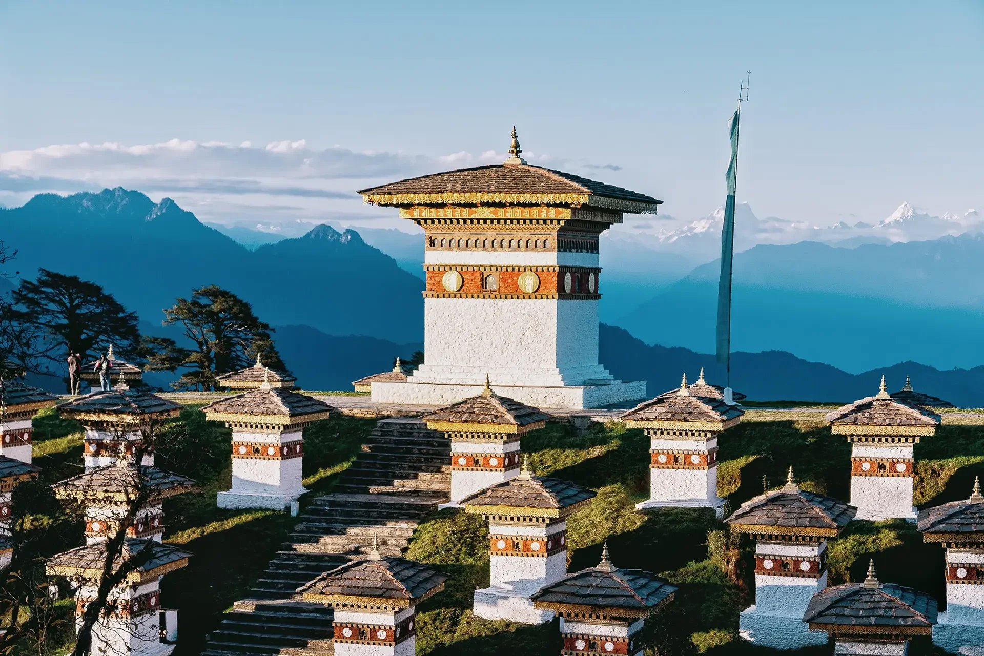 A traditional Bhutanese dzong with a large white stupa in the center, surrounded by smaller stupas, against a backdrop of mountains and a clear blue sky.