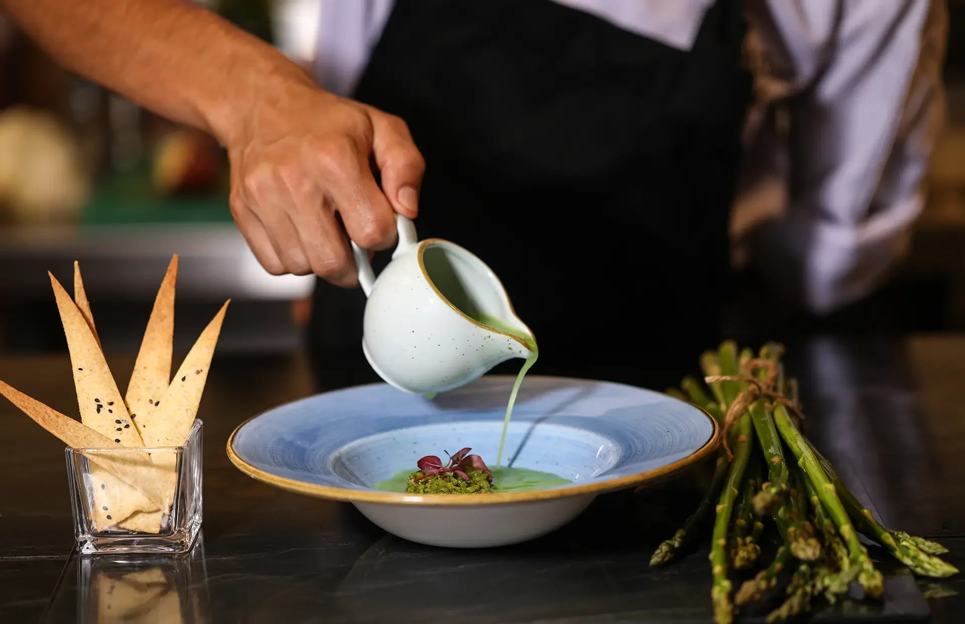 Chef pouring green sauce or soup into a bowl with garnishes, served on a decorative plate, with crispy breadsticks in a glass container and asparagus on the side.