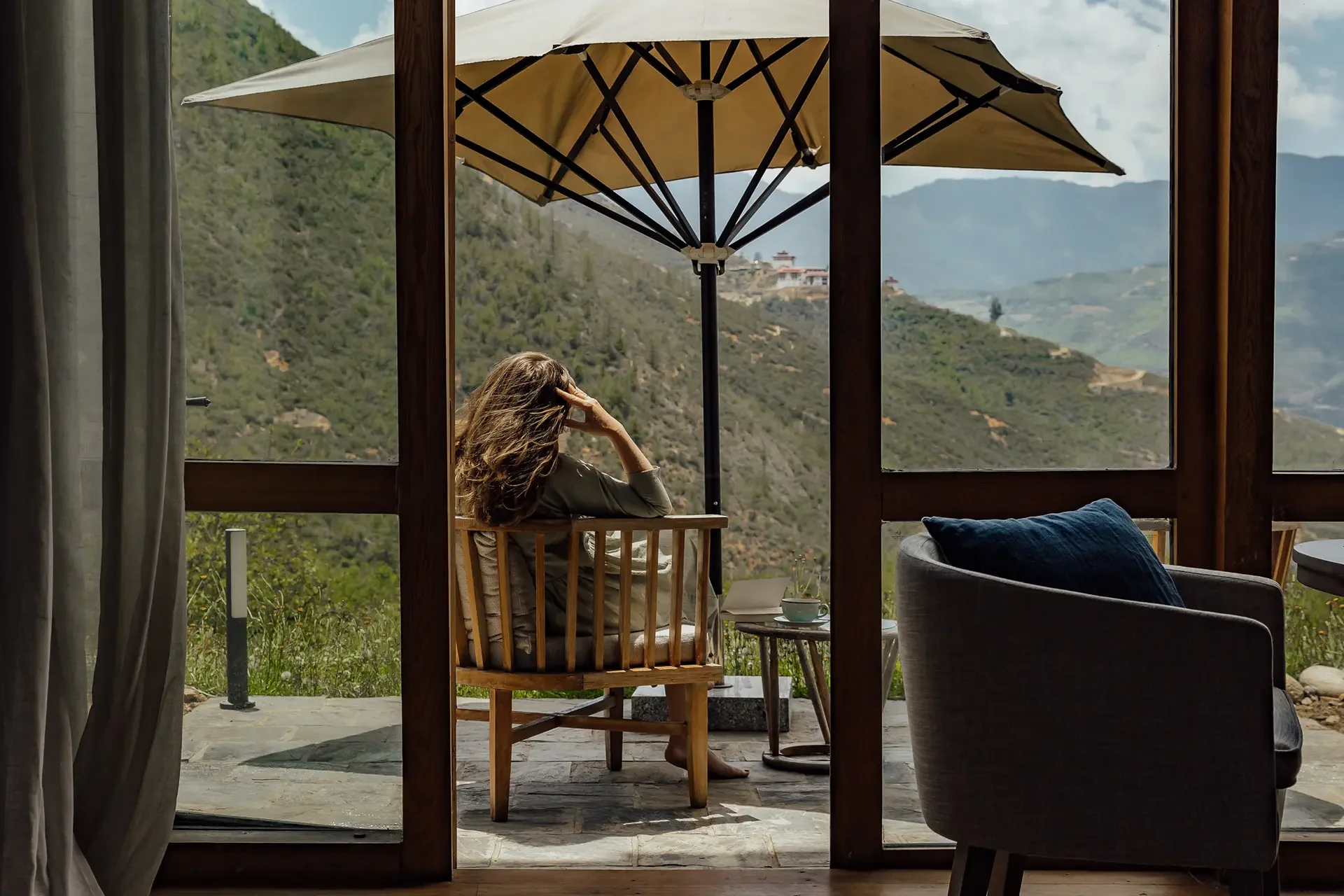 A woman sitting on a wooden chair on a balcony with a mountain view, partially shaded by large beige umbrellas, with a cup and saucer on a small table nearby.