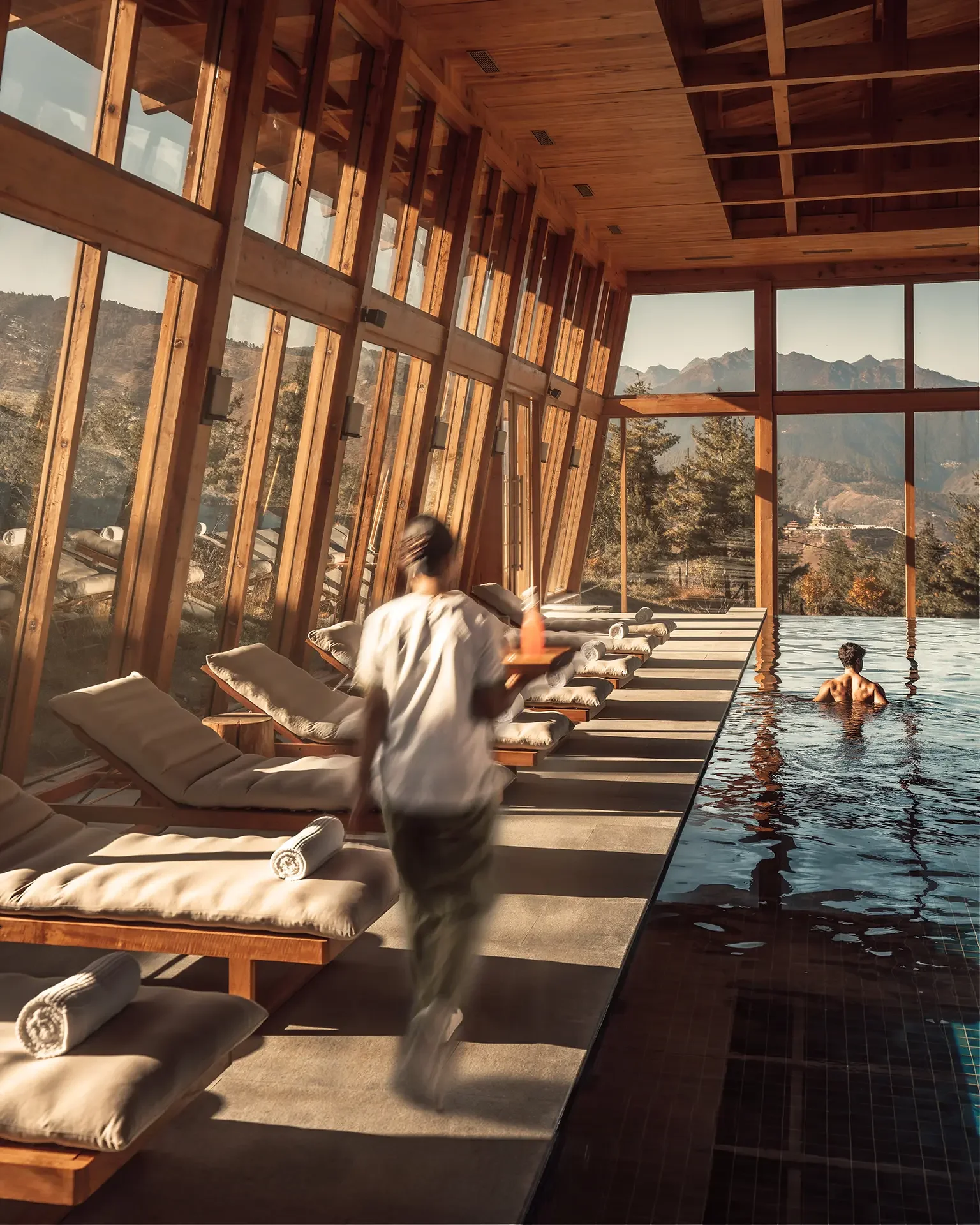 Interior view of a wooden spa or pool area with lounge chairs, a person in the pool, and large windows showing mountains and trees outside.
