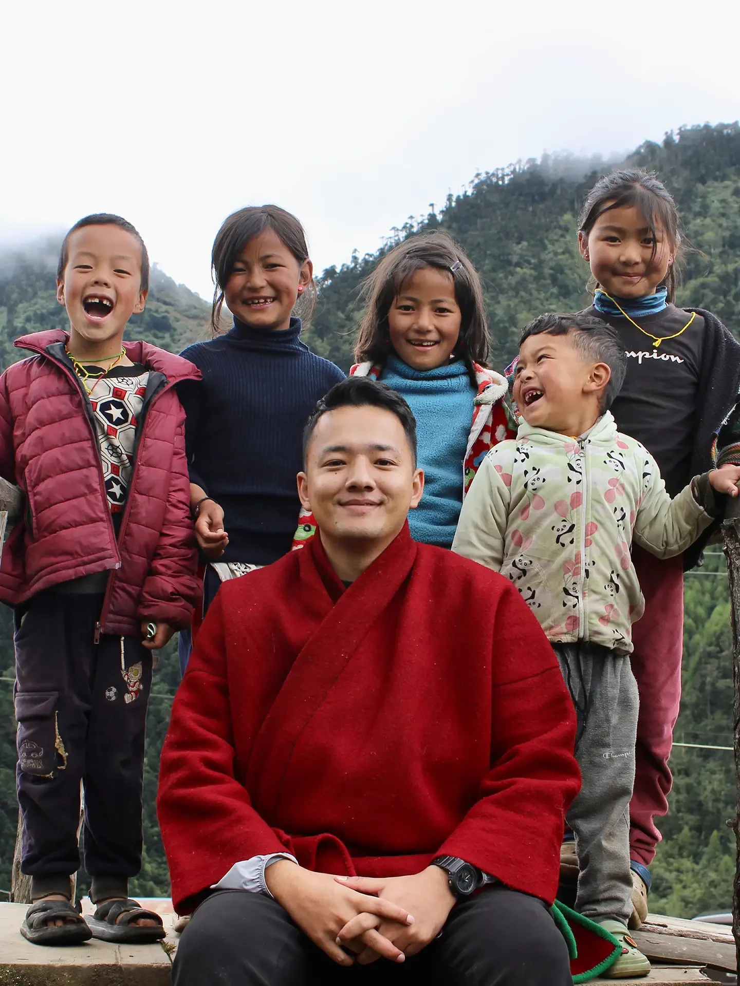 A group of smiling children and a man in a red robe outdoors with a mountain background, some children wearing jackets and one child in a panda print jacket.