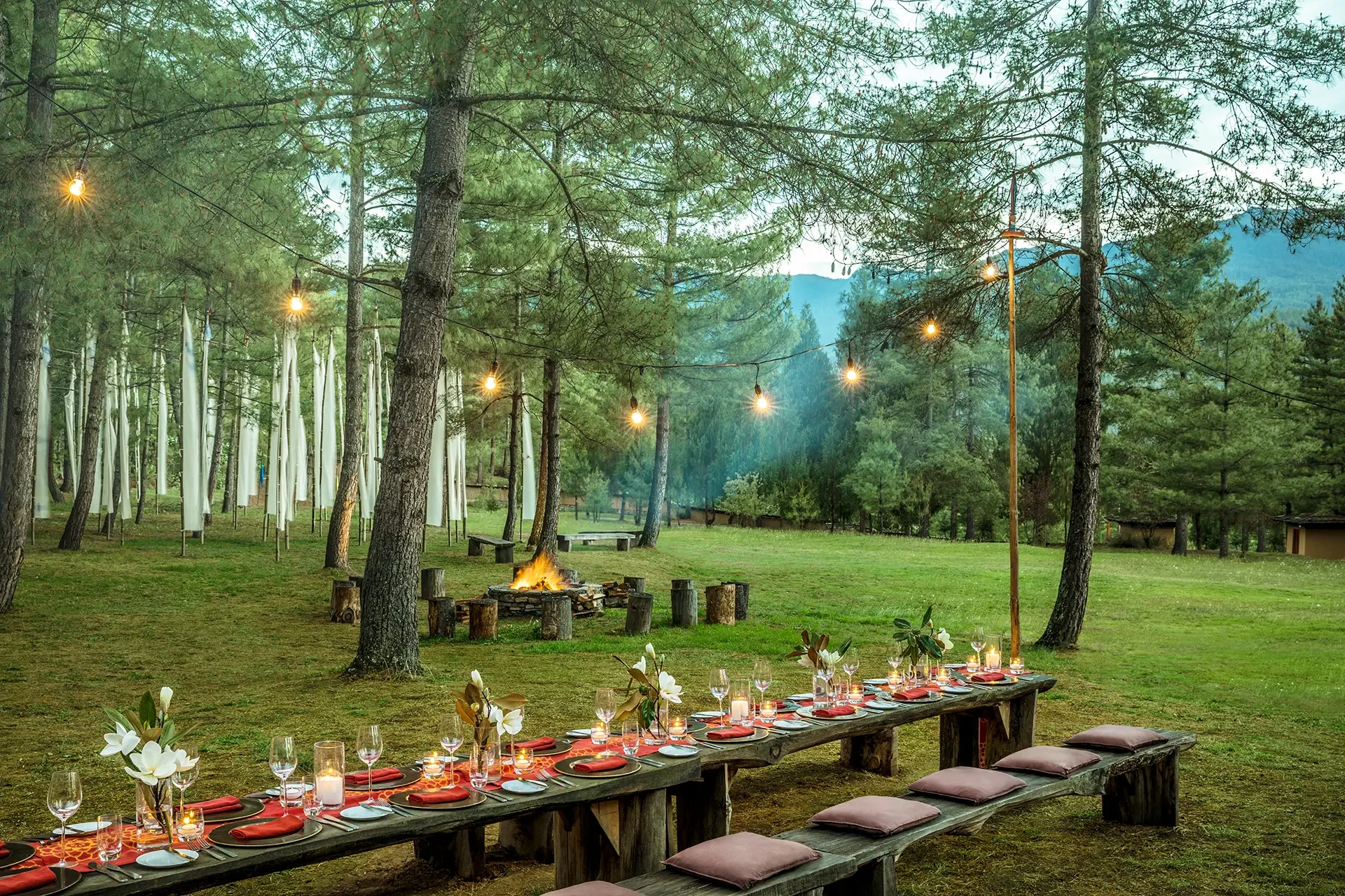 Outdoor dining setup with a long wooden table decorated with flowers, candles, water glasses, and red napkins, set in a forest with string lights, a campfire, and mountains in the background during twilight.