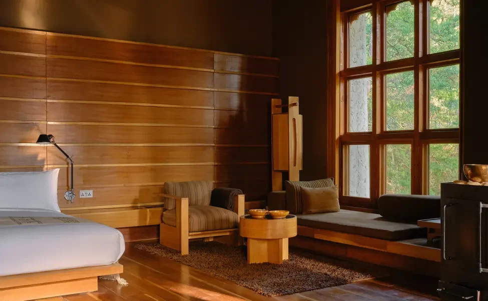 A cozy bedroom corner with a bed, wooden wall paneling, a window seat with cushions, and natural light coming through large window panes.