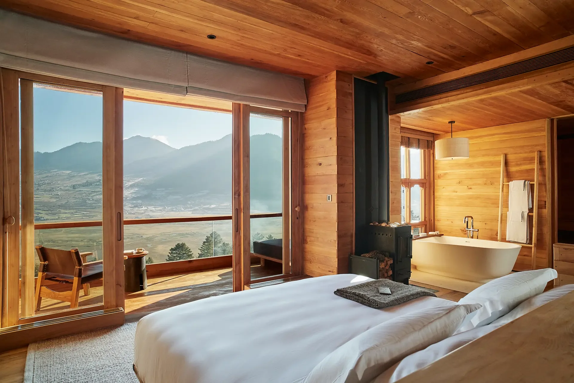 Interior of a cozy wooden cabin bedroom with large windows showing a mountain view, a bathtub, a wooden ladder with a towel, and a small sitting area.
