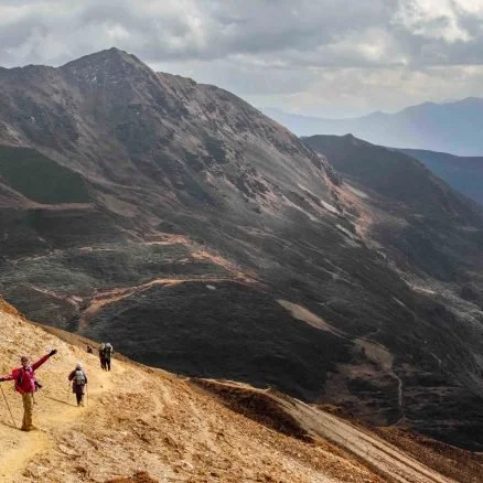 Hikers walking along a mountain trail with rugged hills and cloudy sky in the background.