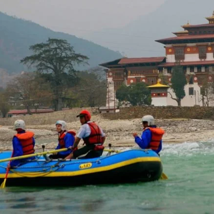A group of four people wearing life jackets and helmets are rafting on a river with a traditional Nepalese-style building and mountains in the background.