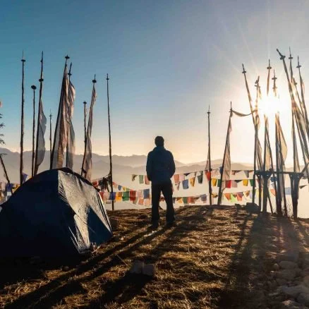 A person standing outdoors at sunrise or sunset near a small tent and prayer flags, with sailboat masts in the background.