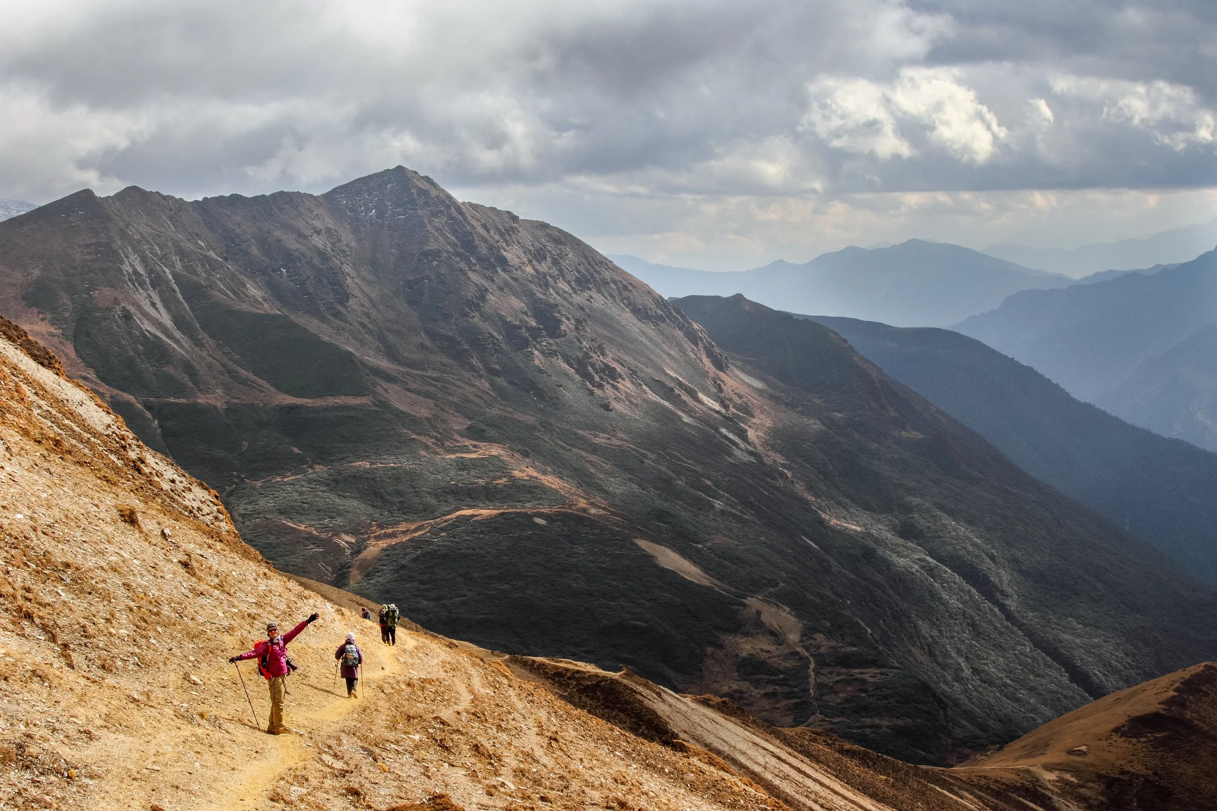 Hikers on a mountain trail surrounded by rugged, rolling mountains under cloudy skies.