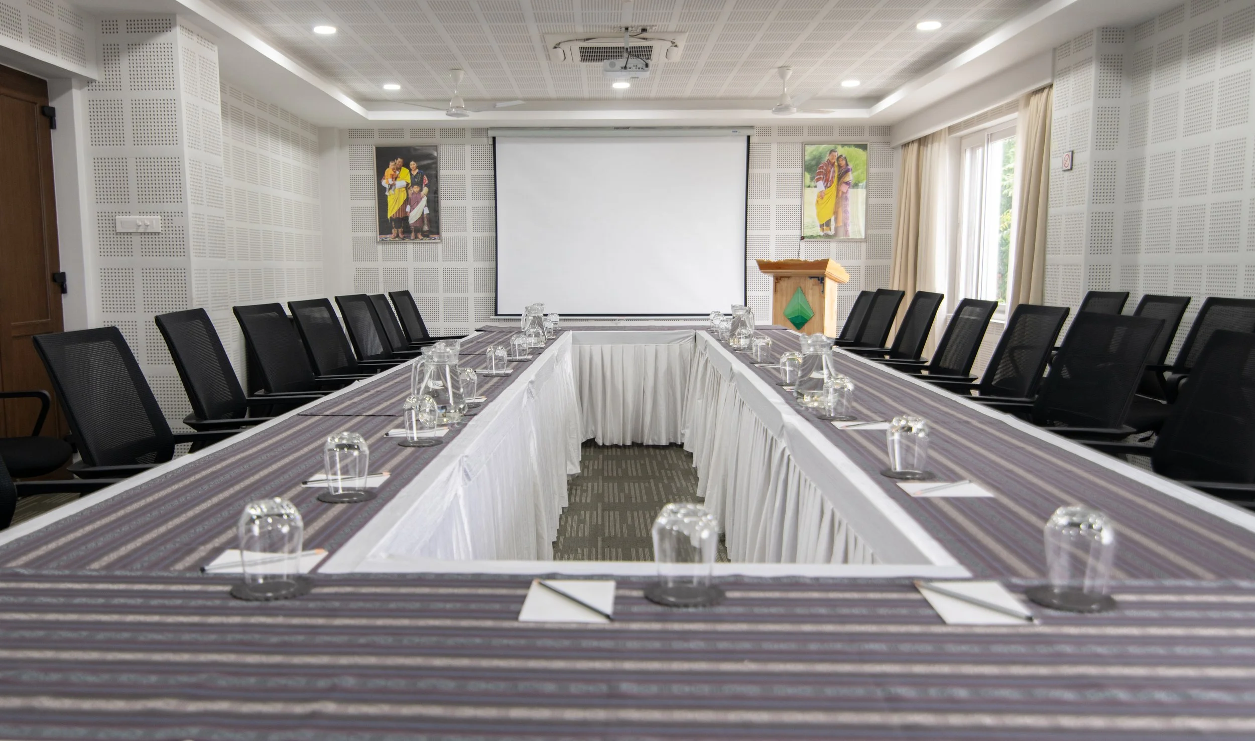 Conference room with U-shaped table setup, black mesh chairs, and glass water glasses, with a white projection screen at the front and portraits on the walls.
