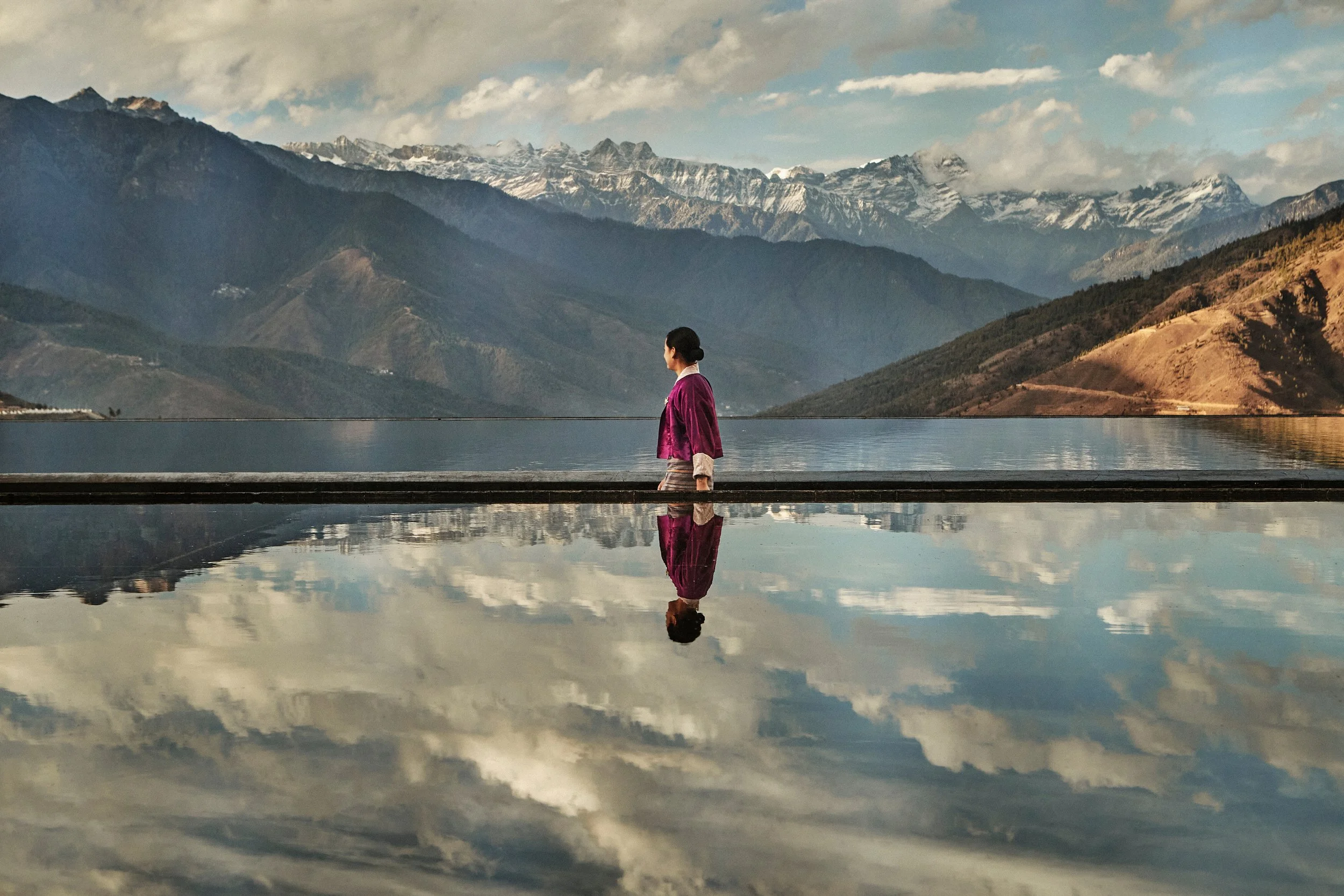 A woman in a purple jacket stands on a platform near a body of water, with mountains in the background and their reflection visible in the water.