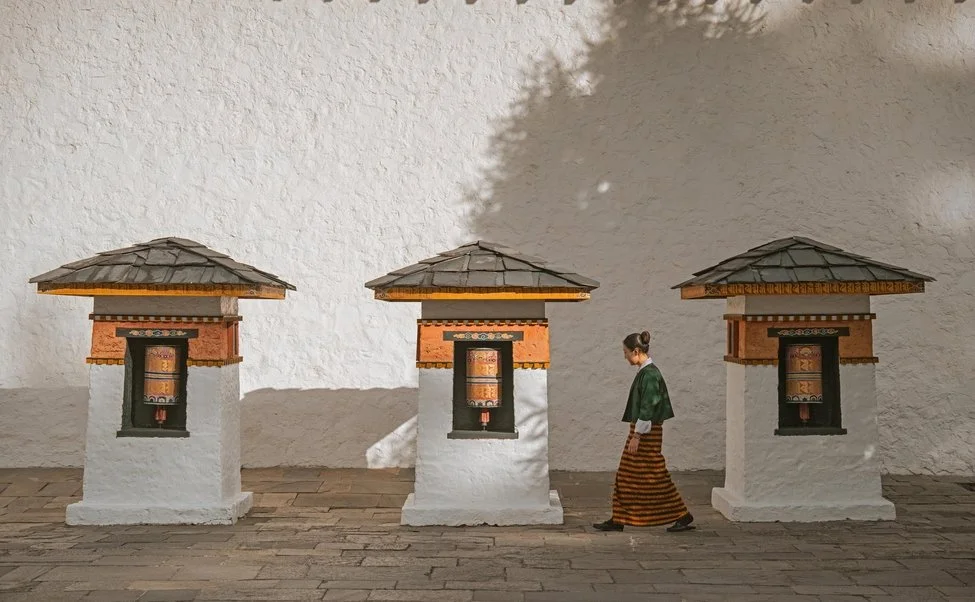 A woman in traditional Bhutanese clothing walking past three small white pagoda-style structures against a textured beige wall.