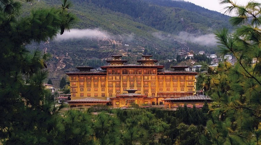 A large, traditional-style building, possibly a hotel or palace, situated among lush greenery with mountain hills in the background.