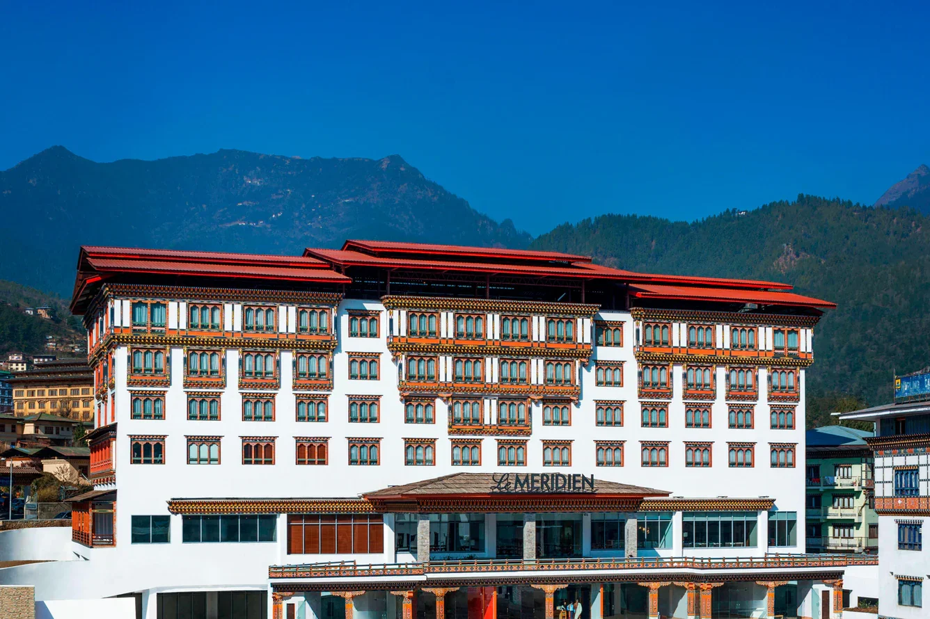 Front view of a large white hotel building with traditional Bhutanese architecture, including decorated windows and a red roof, set against green mountains and a clear blue sky.