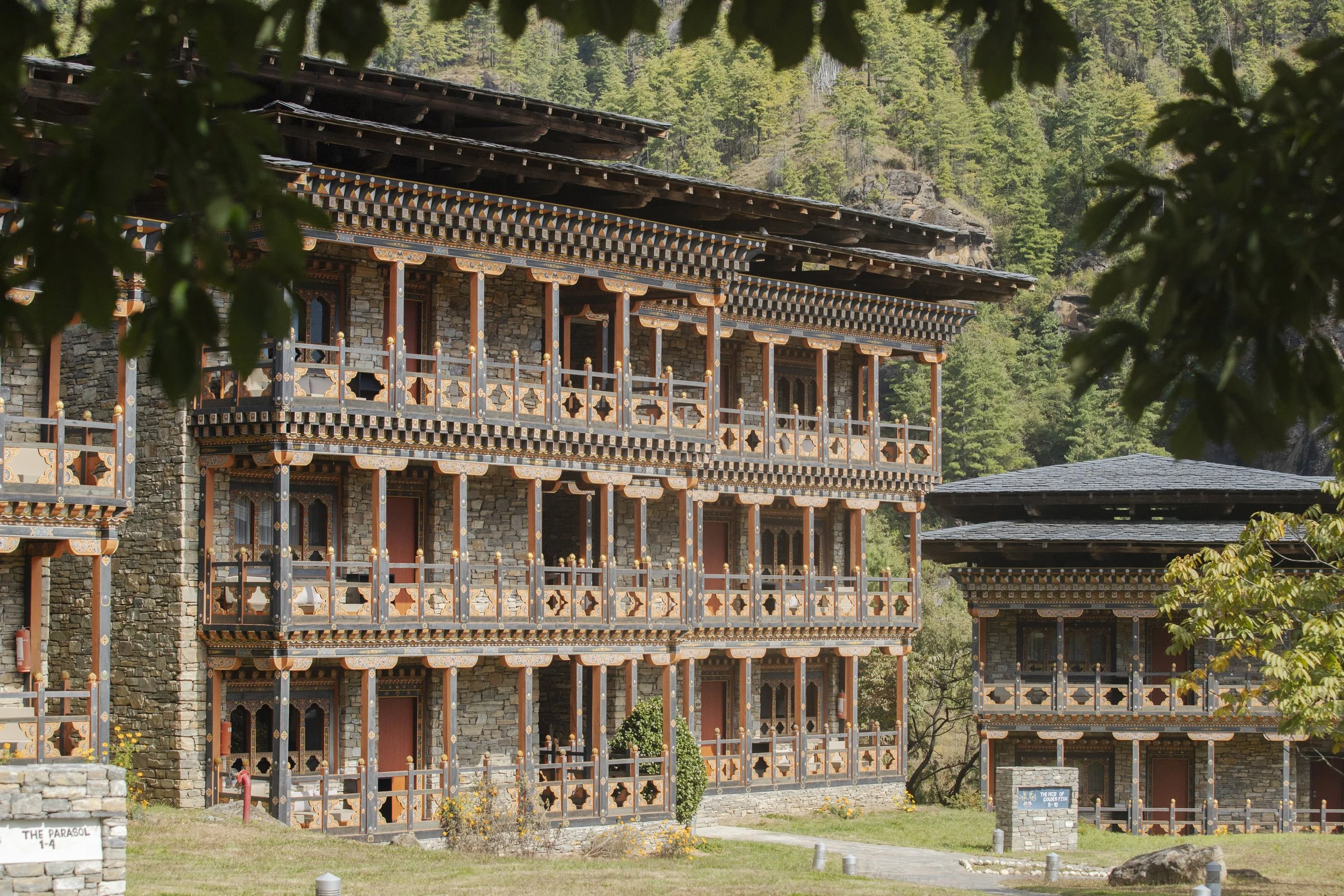 Traditional multi-story wooden building with intricate architectural details, set against a backdrop of green trees and mountains, partially framed by branches and leaves.