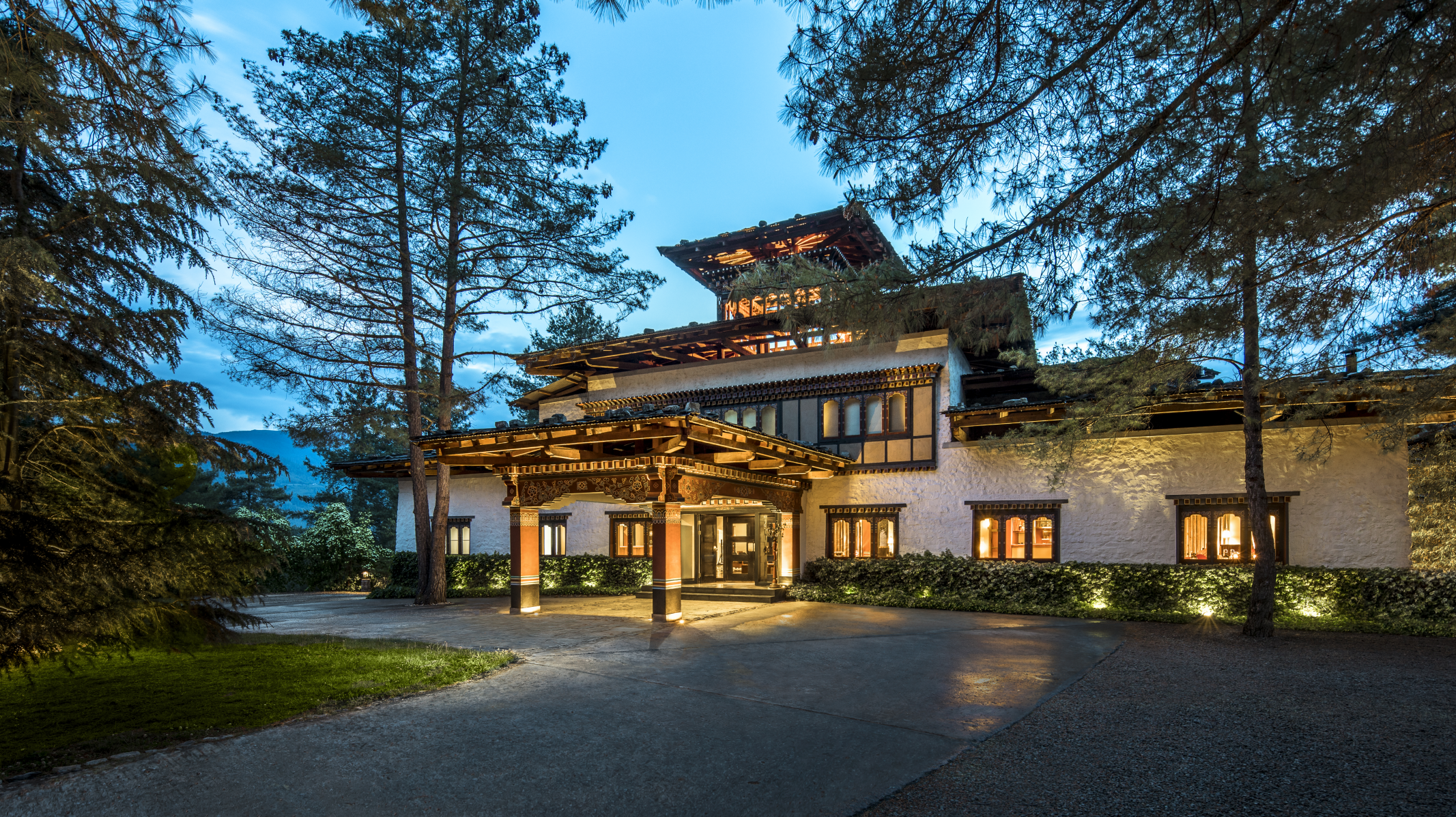 A large house with traditional architectural features, surrounded by tall trees and illuminated at night, with a paved pathway in the front.