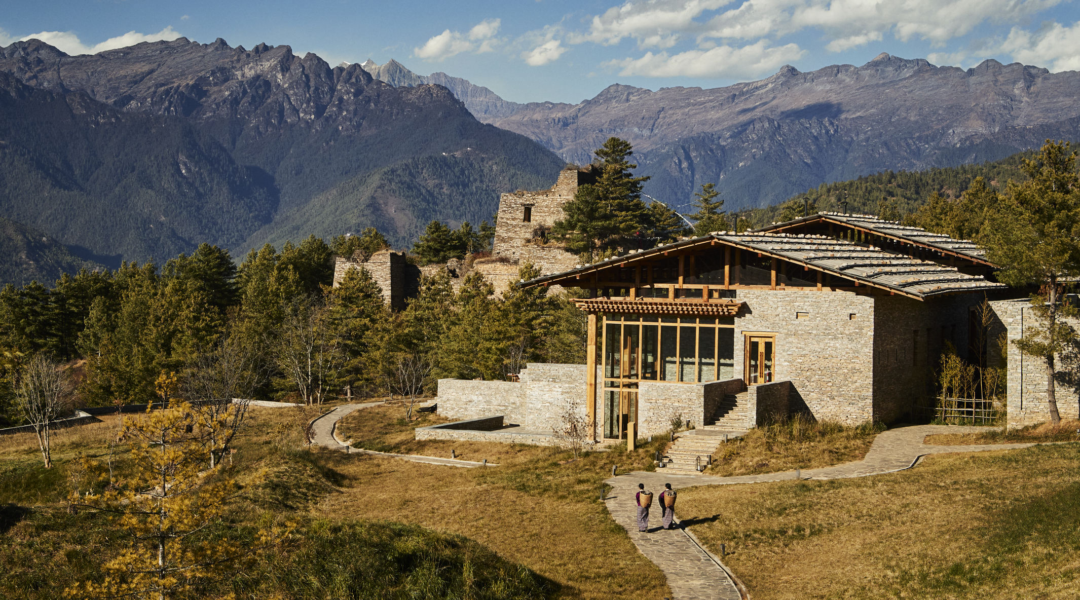 Modern stone house with large glass windows and wooden accents, surrounded by trees and mountains in the background, with a pathway leading to the house and two people walking.