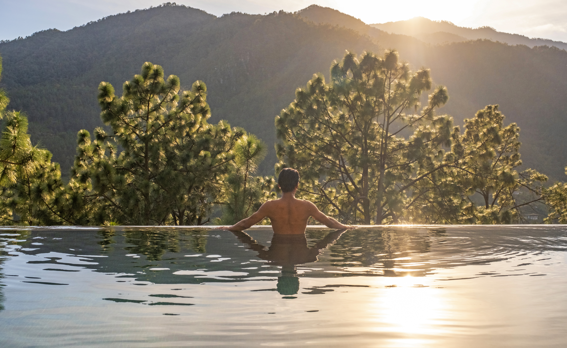 A person bathing in an infinity pool with a natural landscape of trees and mountains at sunset or sunrise.