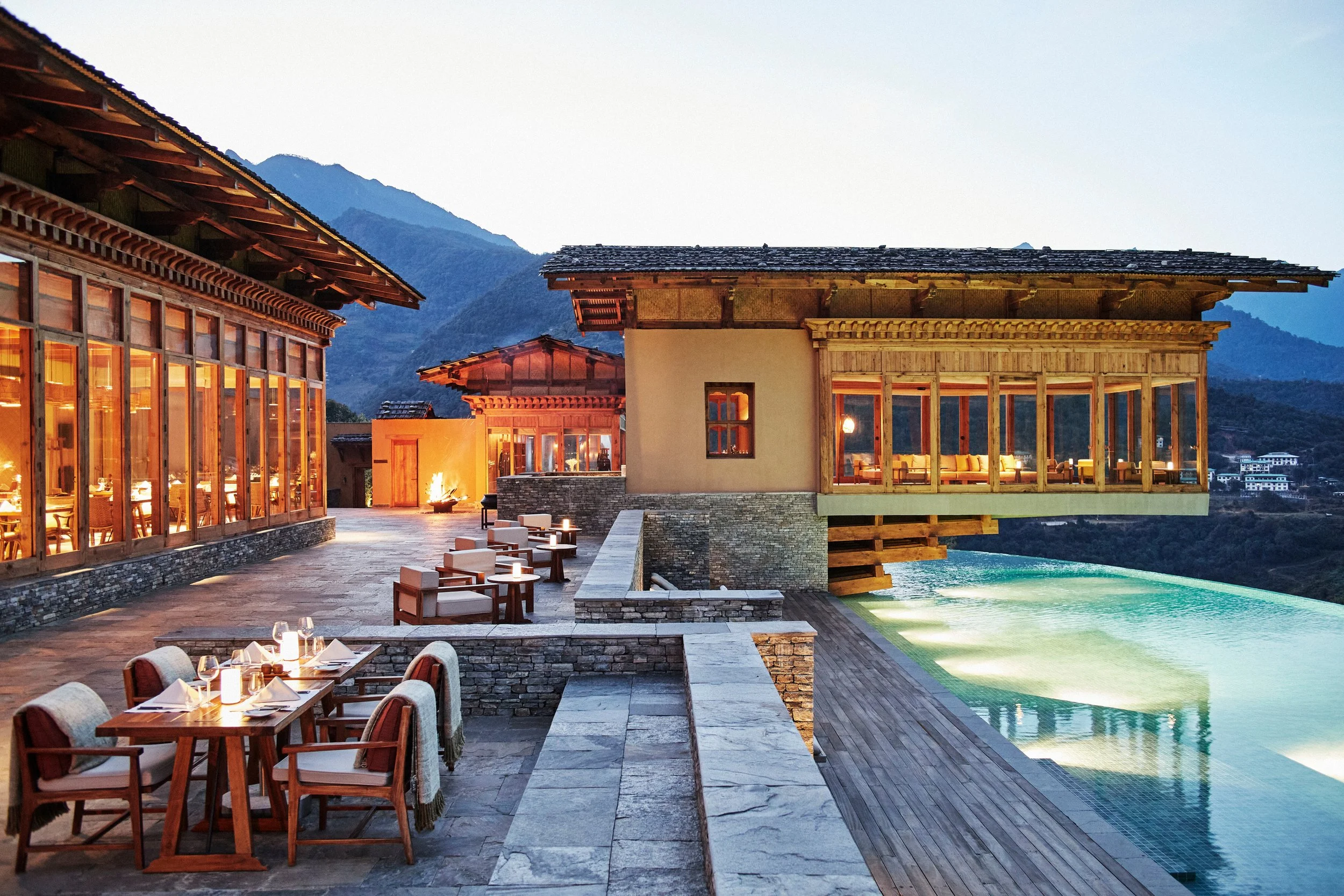Luxury outdoor restaurant with wooden tables and chairs, lit with candles, overlooking a pool with mountain scenery in the background during evening.