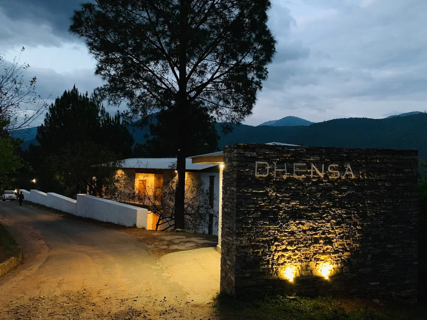 Entrée area of a building with a large stone sign that reads 'DLIENSA' illuminated by ground lights, surrounded by trees and mountains in the background during dusk or early evening.
