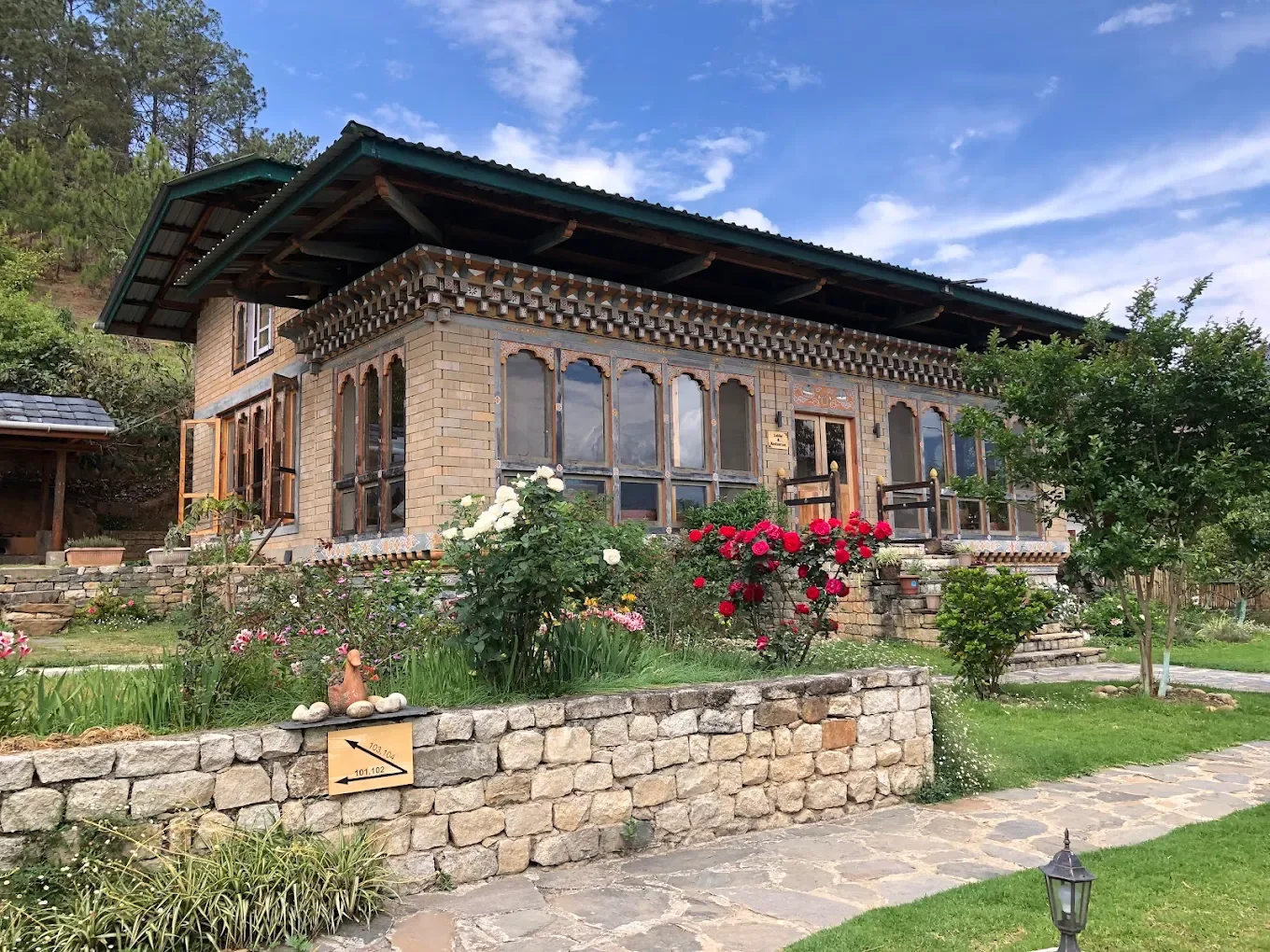 A house with brick and wood exterior, large windows, and a garden with flowers, bushes, and a stone pathway leading to the entrance, under a partly cloudy sky.