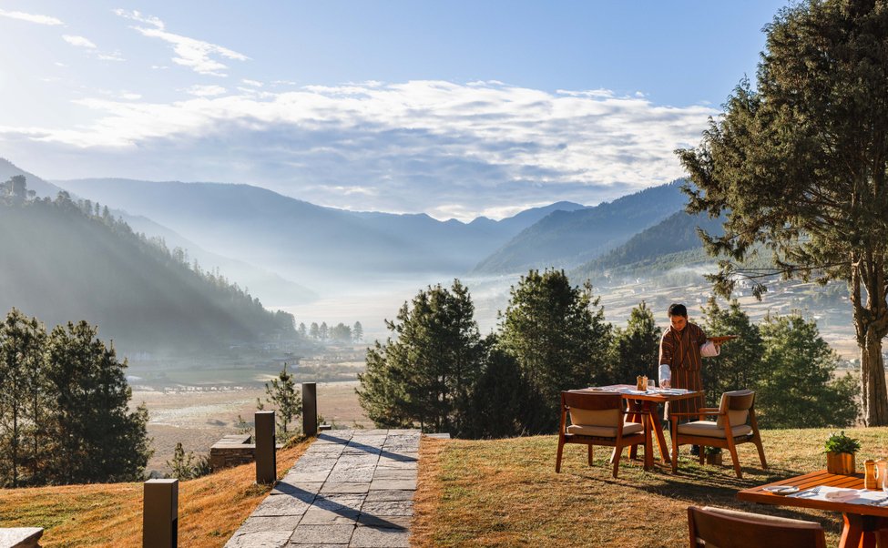 A person setting a table outdoors on a grassy area with a scenic mountain landscape in the background.