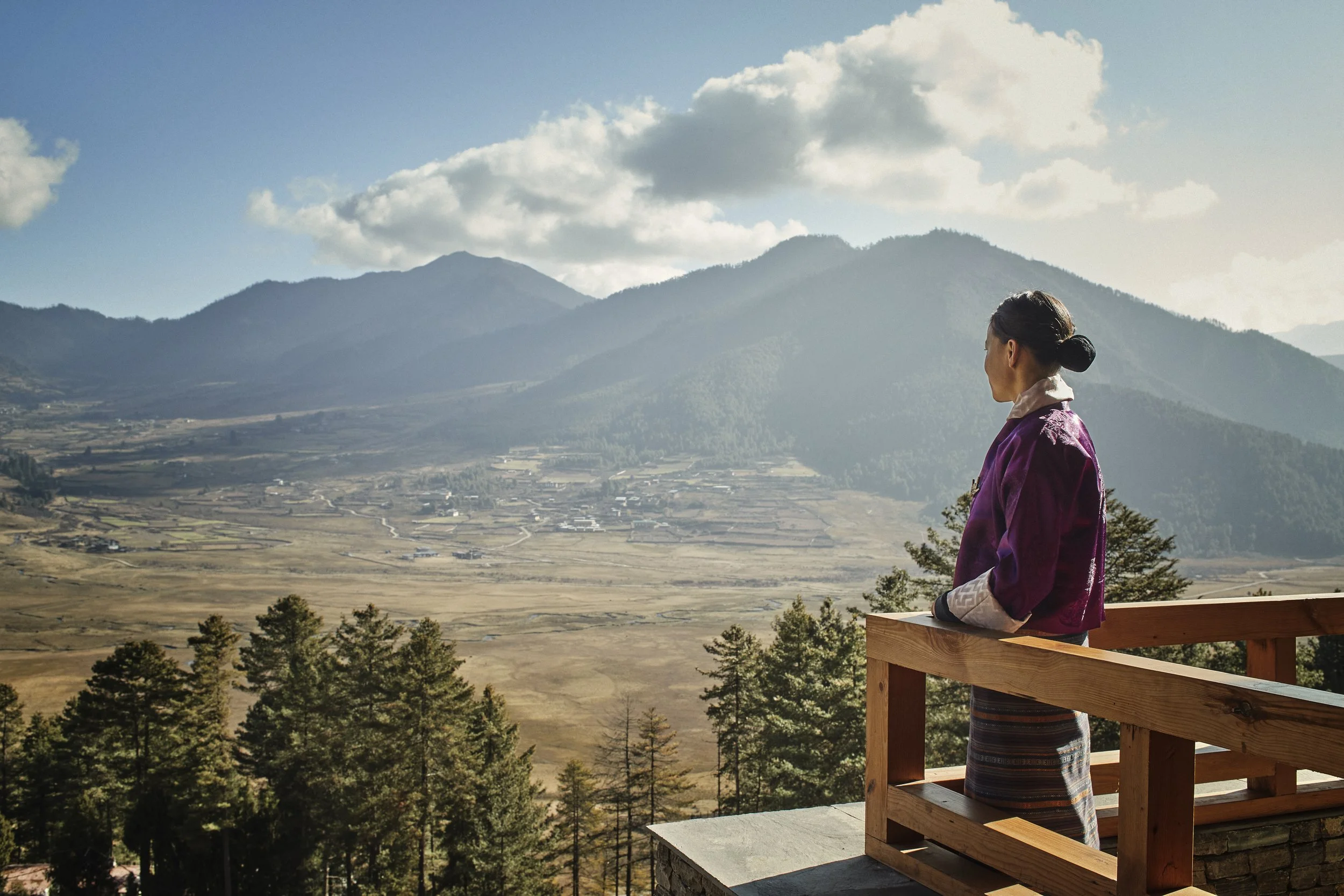 A woman wearing traditional Asian clothing standing on a balcony overlooking a valley with mountains and cloudy sky.