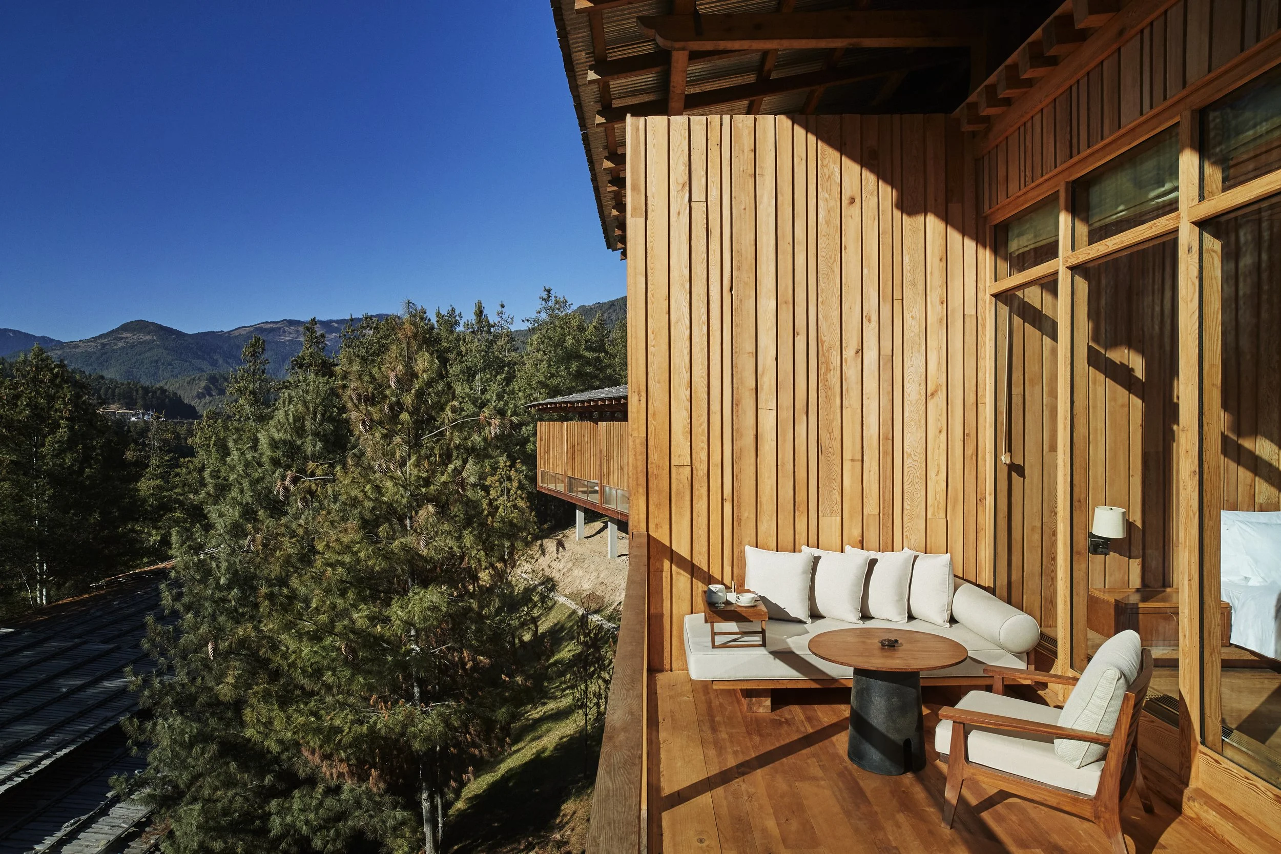 Balcony with outdoor seating area featuring a white sofa, armchair, and small table, overlooking lush green trees and mountains on a clear, sunny day.