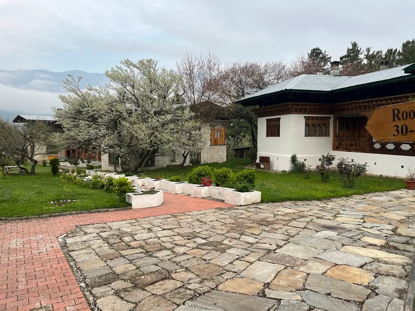 Scenic view of a traditional house with wooden details, surrounded by blooming trees and manicured garden, with a cloudy sky in the background.