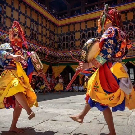 Two dancers dressed in colorful traditional costumes perform a dance in an open courtyard, with a decorated cultural building in the background.